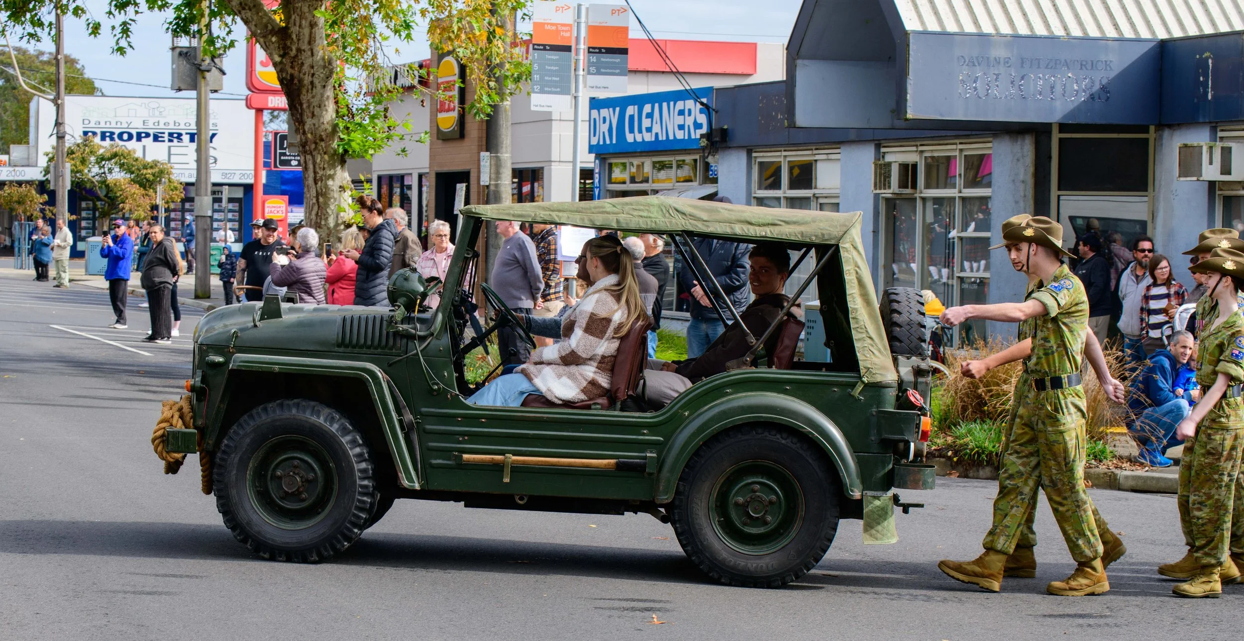 A military-style green Jeep with two seated women, one wearing a plaid coat, being led by three women in army fatigues during a parade on a city street with shops and spectators.