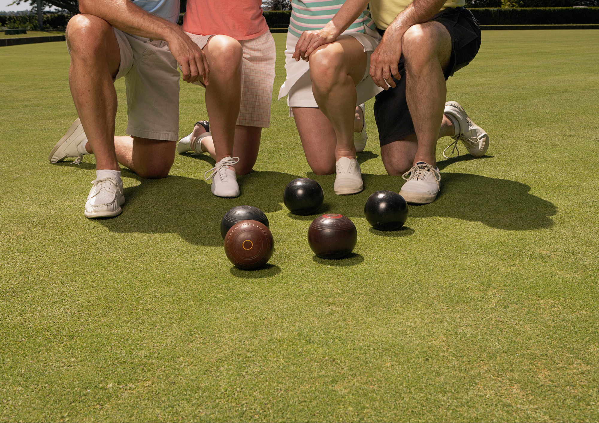Four people kneeling on a putting green, preparing to play lawn games with black and brown bocce balls.