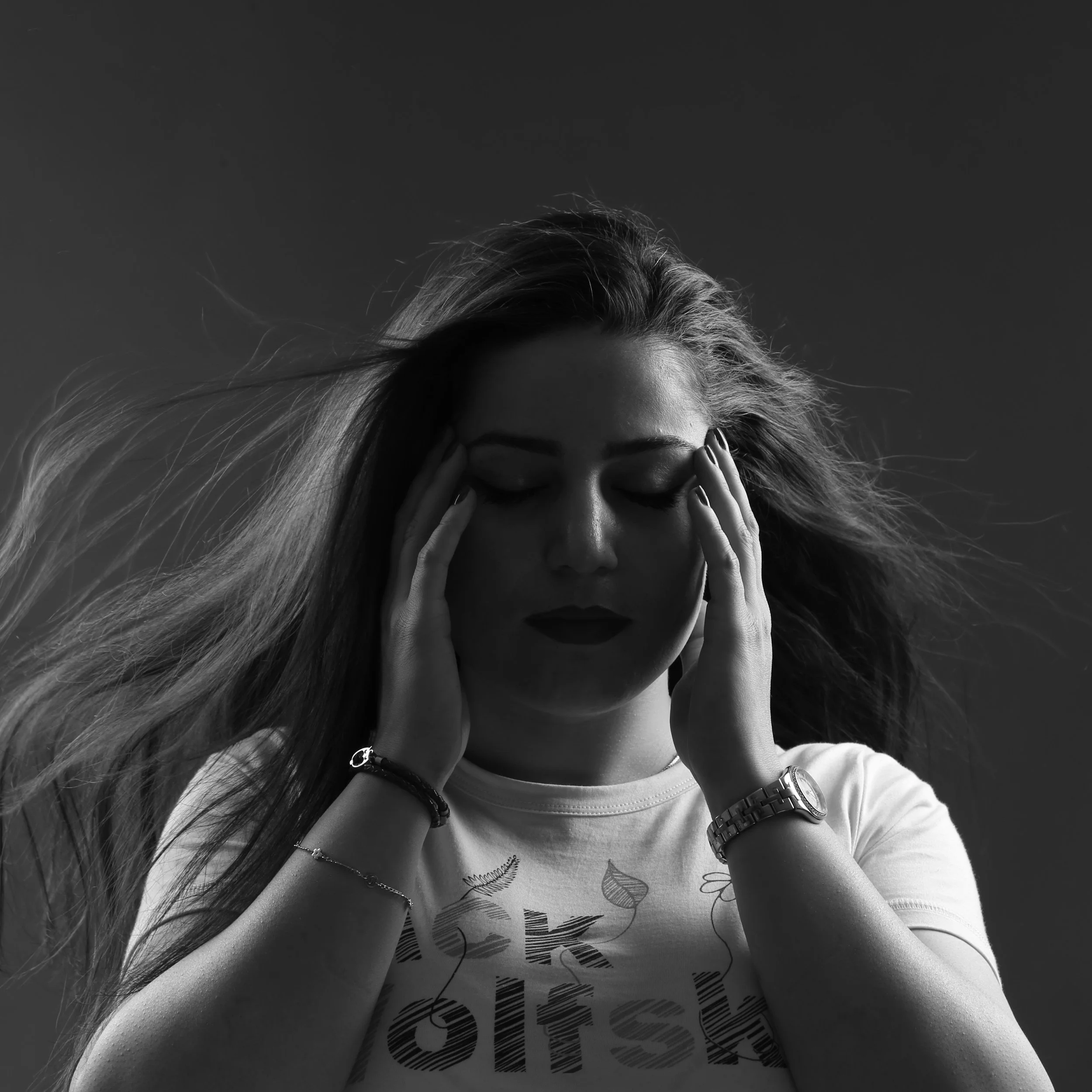 A black and white photograph of a woman with long hair covering part of her face, wearing glasses and a wristwatch, with her hands on her head, against a cloudy sky background.