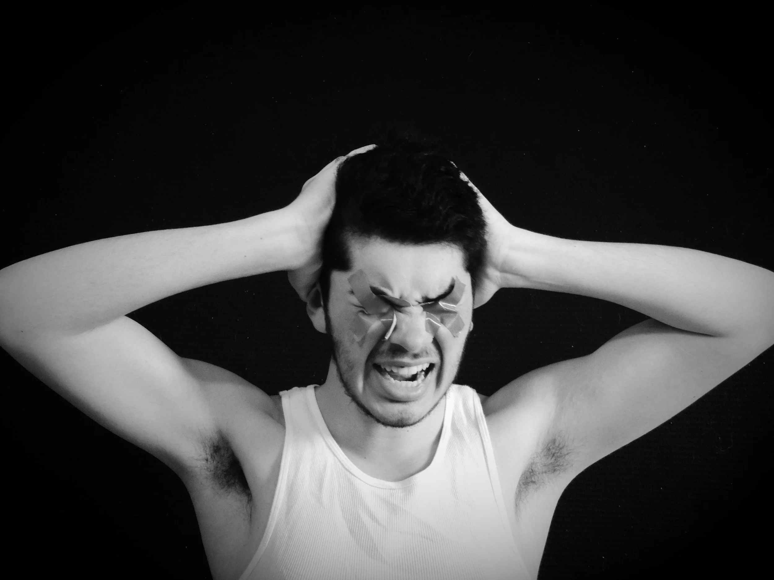 A person with short hair, wearing a sleeveless shirt, laughing while holding their head with both hands, eyes closed, against a black background.