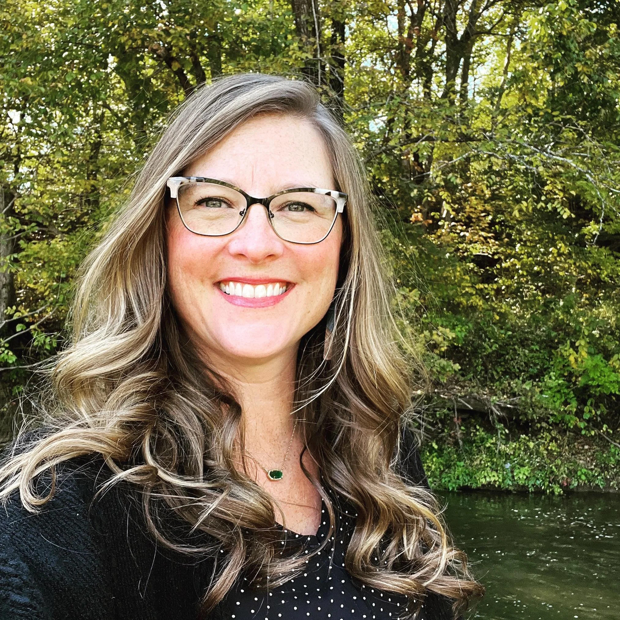 Close-up of a woman with glasses and long, wavy blonde hair smiling outdoors, with trees and foliage in the background.