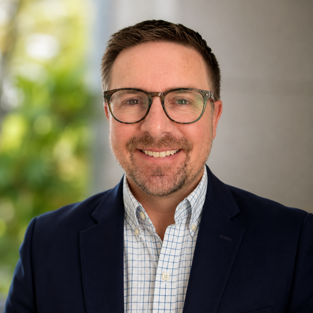 Professional headshot of a smiling man wearing glasses, a navy blazer, and a checkered button-up shirt, with a blurred green and light background.