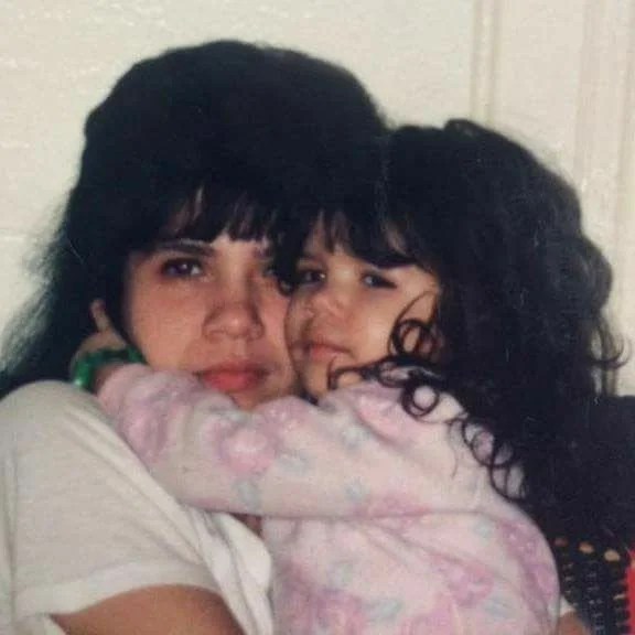Two young girls, one older and one younger, hugging each other closely and looking at the camera. Both have dark, curly hair.