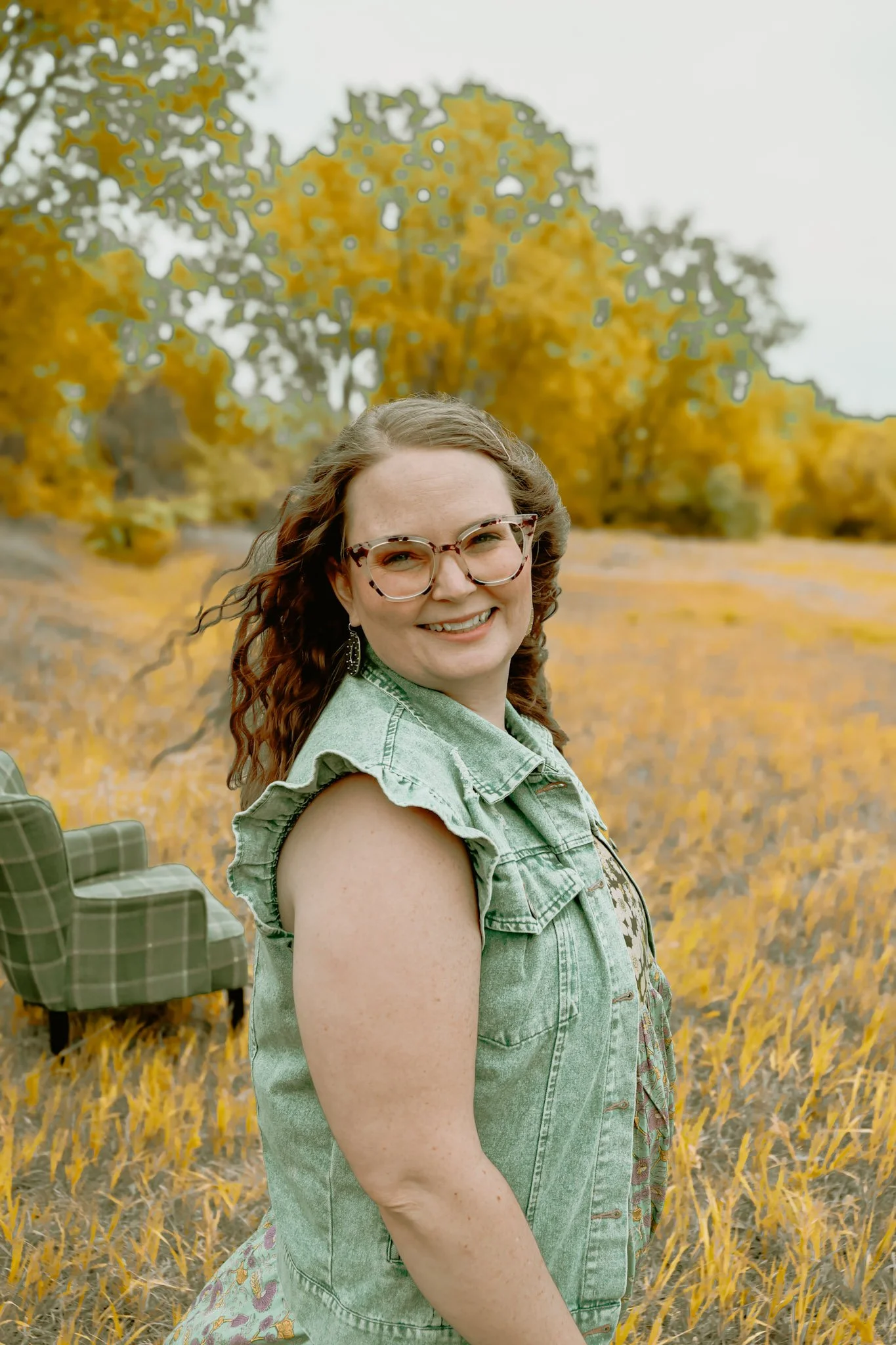 A smiling woman with curly hair and glasses standing in a field of yellow flowers during autumn, with trees in the background.