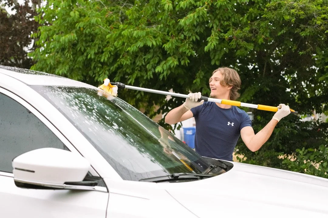 A person cleaning a white car with a long-handled squeegee, standing outdoors near green trees.