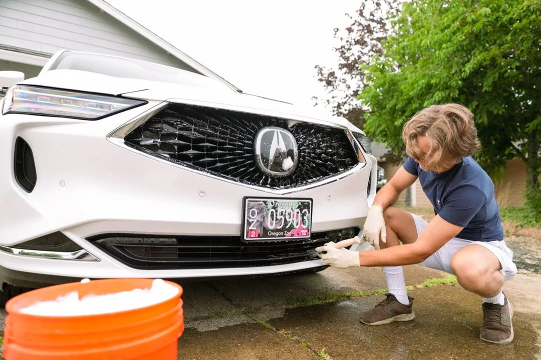 A person with blonde hair, wearing a navy blue shirt, white shorts, and gloves, is crouching down and cleaning the front license plate of a white Acura car parked outside near trees and a house.