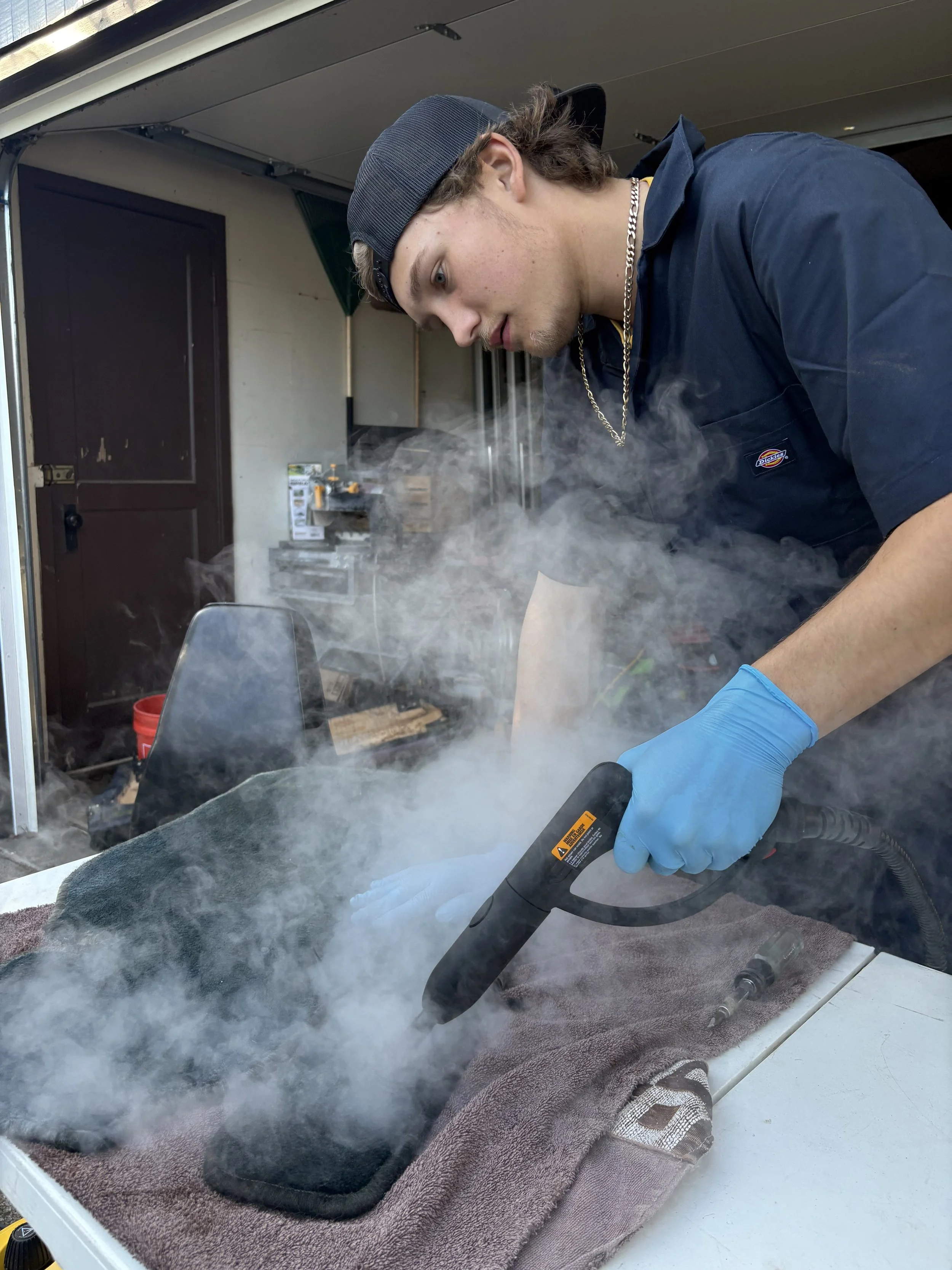 A person using a handheld steam cleaner on a black object covered with a brown towel. The person is wearing a blue uniform, a chain necklace, a black cap, and blue gloves. Steam is visible around the object, and the background includes a door and some storage items.