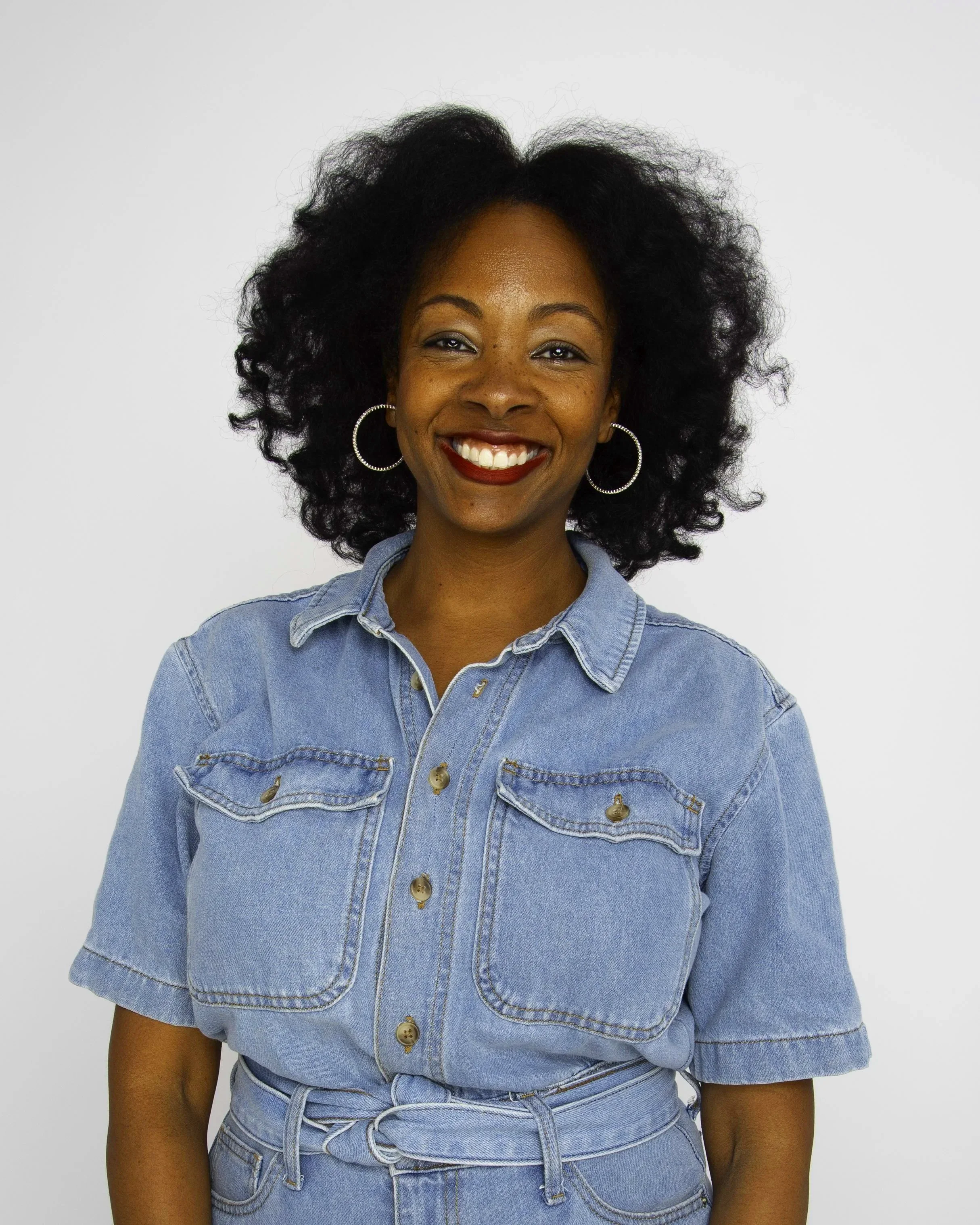 A woman smiling with curly black hair, wearing hoop earrings and a denim shirt against a plain white background.