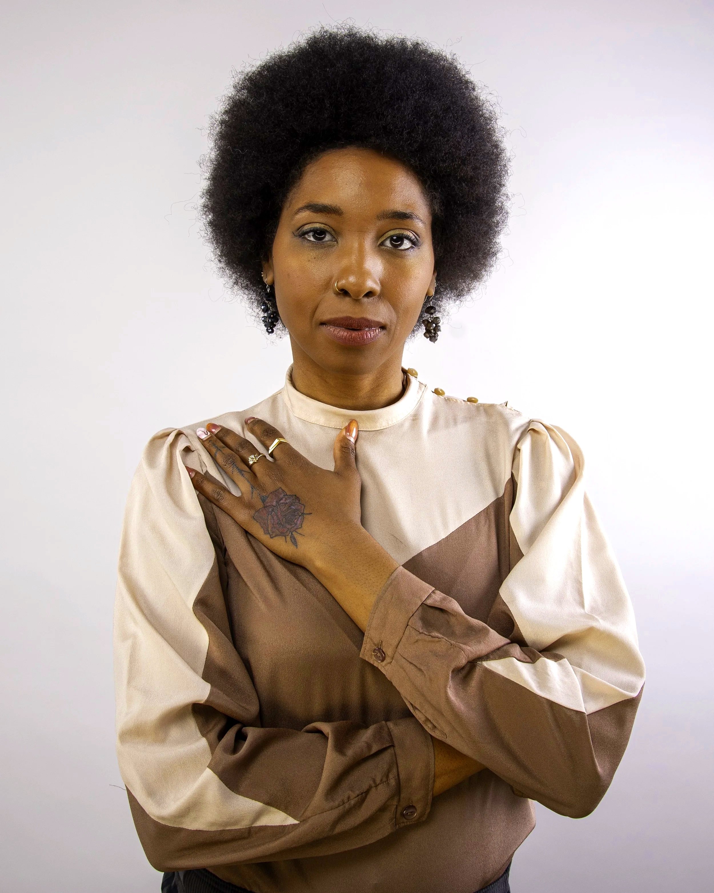 A woman with dark skin and an afro hairstyle posing against a plain white background. She is wearing gold earrings, a nose ring, and a beige and brown color-blocked blouse. Her right hand rests on her chest, showing rings and a floral tattoo on her f