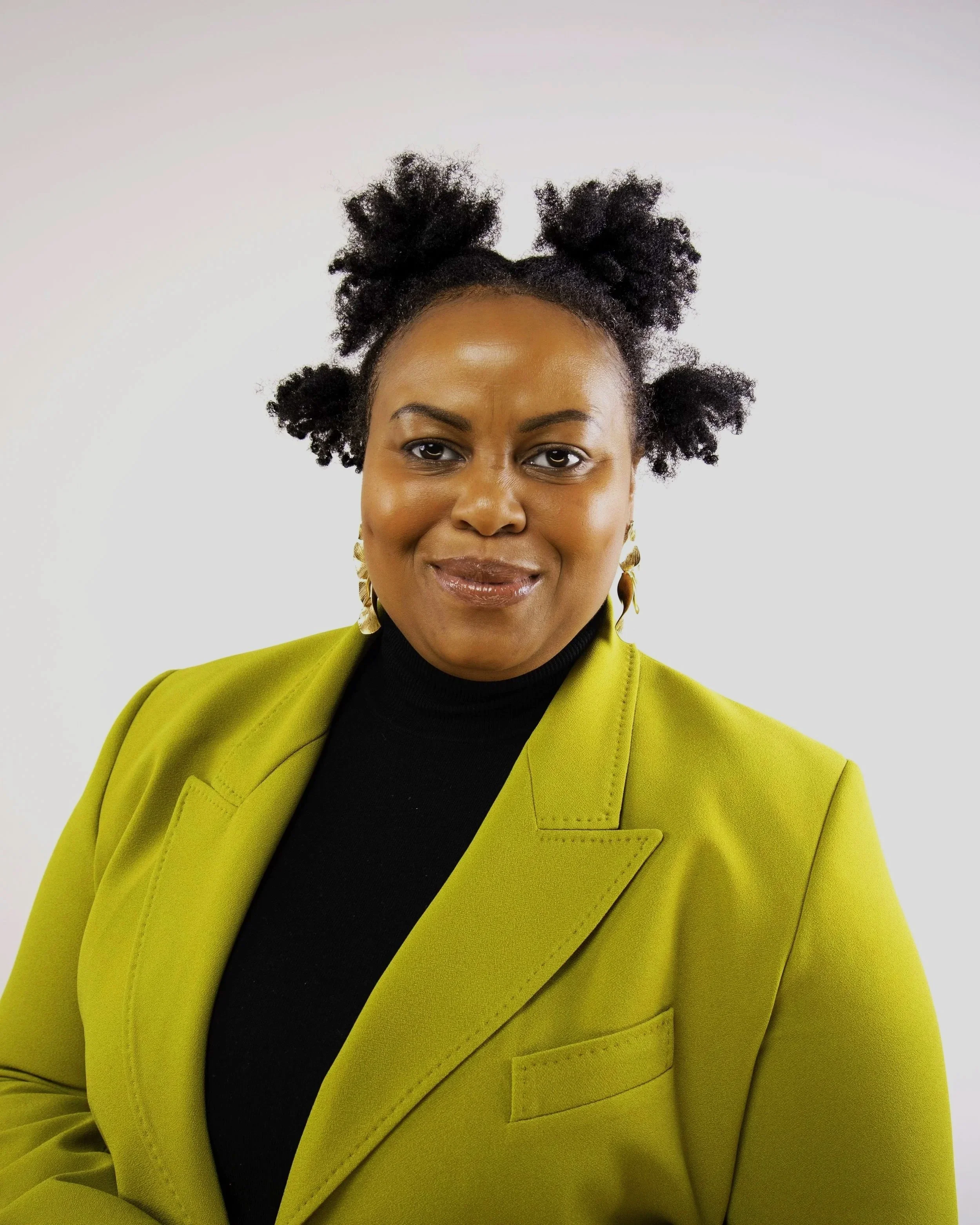 Close-up of a woman with natural hair styled in afro puffs, wearing gold earrings, a black top, and a lime green blazer, against a plain white background.