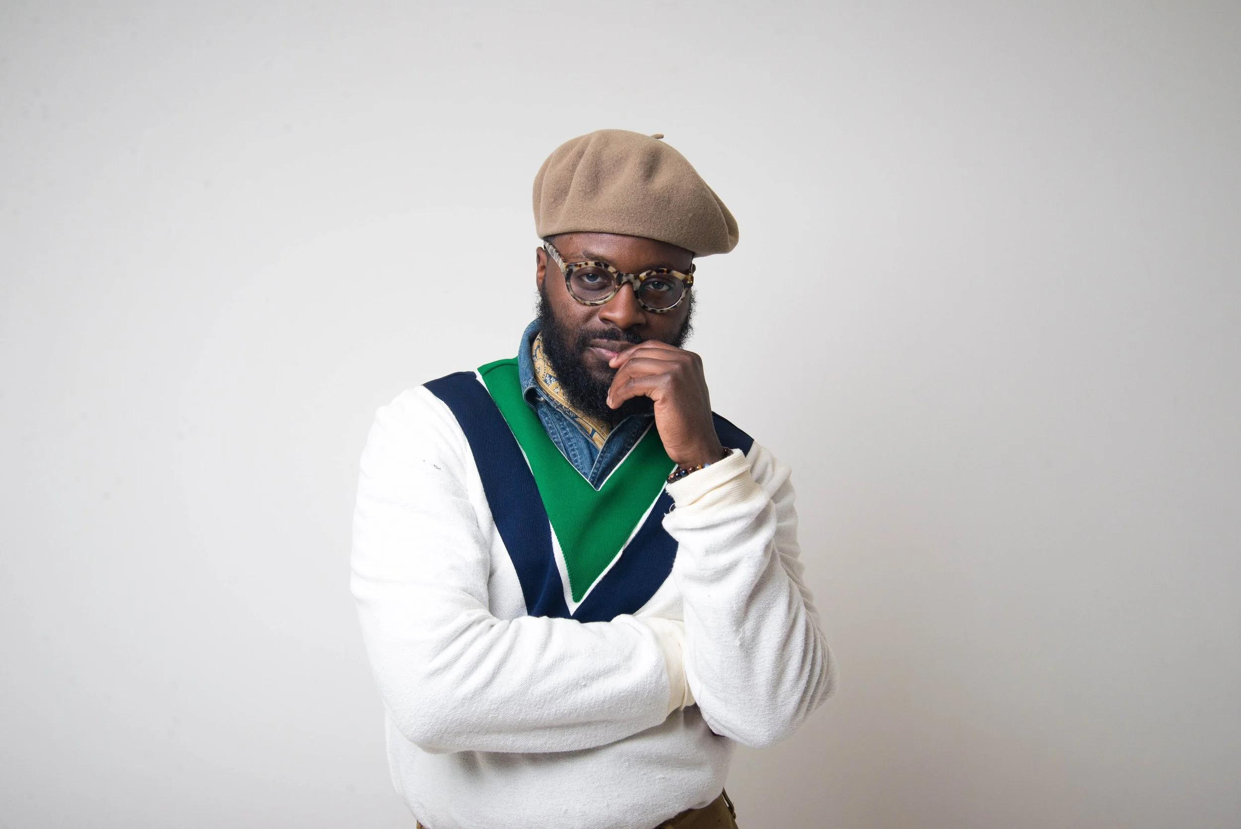 A man with a beard wearing a beige beret, patterned glasses, a denim shirt, a multicolored scarf, and a white sweater with green, blue, and black stripes, posing with his hand near his face against a plain light gray background.