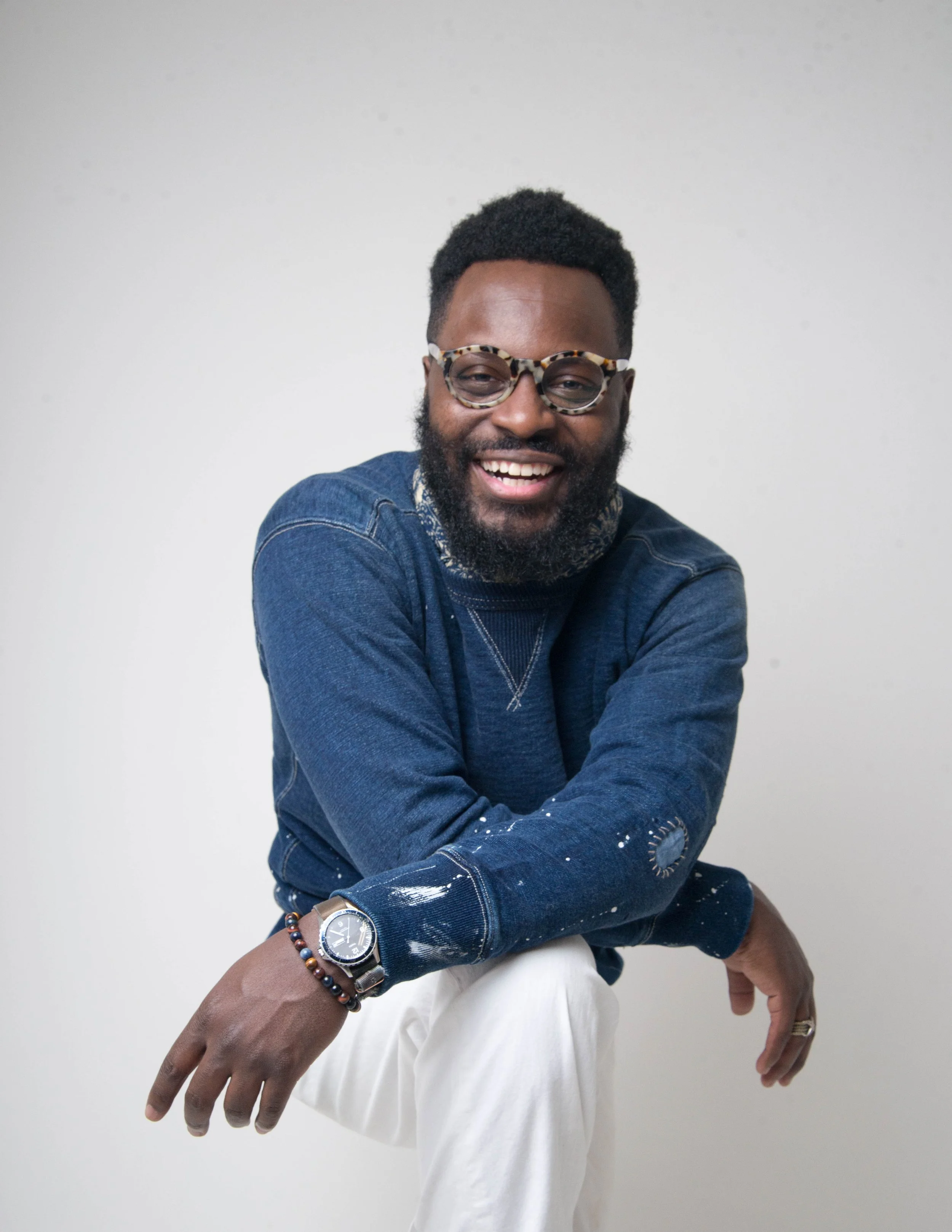 A man smiling and wearing glasses, a denim jacket, a watch, and beaded bracelets, sitting against a plain white background.