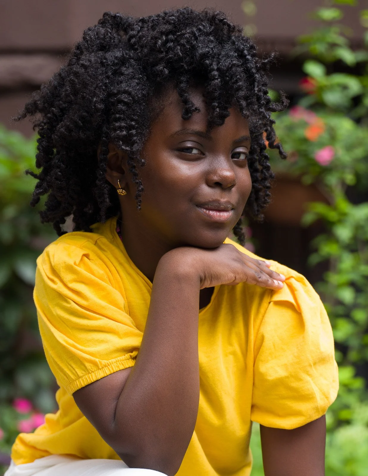 A young woman with dark skin and curly black hair sitting outdoors in front of greenery and flowers, wearing a yellow top, resting her chin on her hand and looking at the camera with a slight smile.