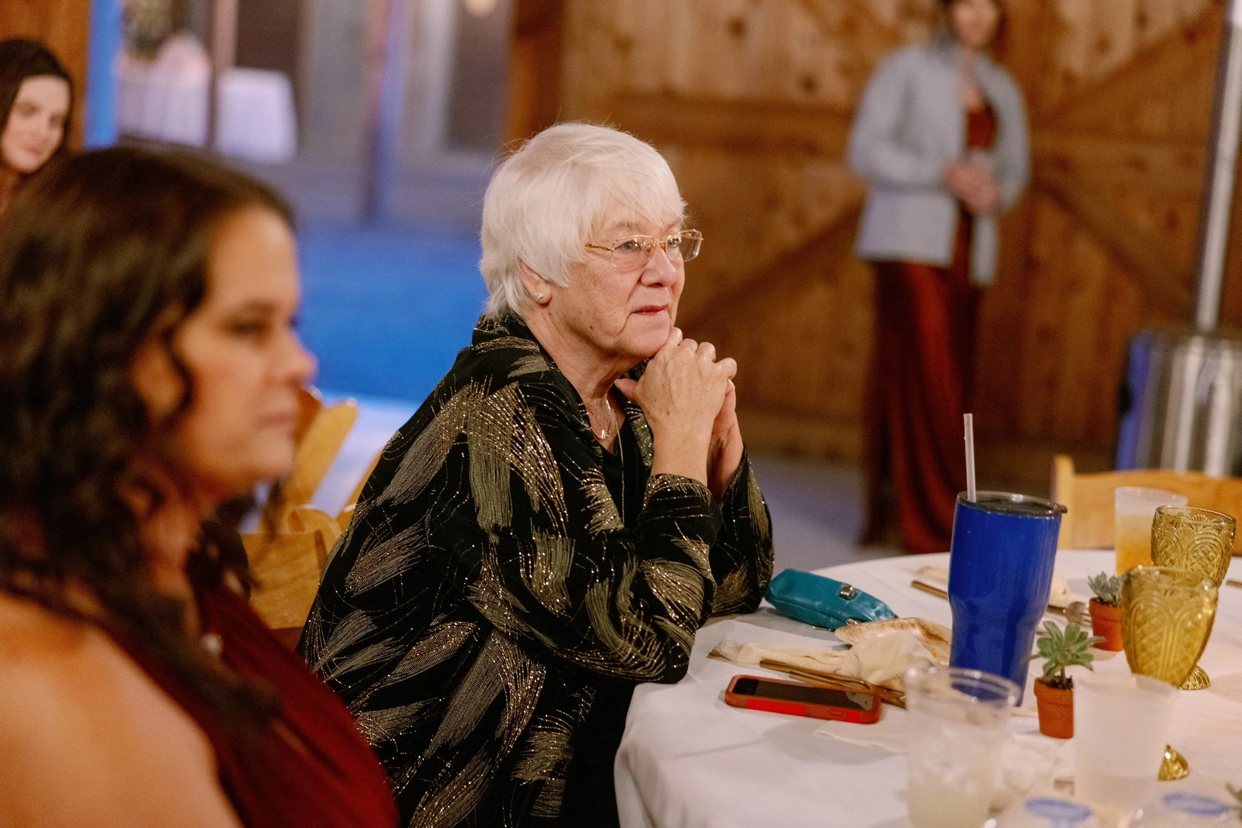 An elderly woman with white hair and glasses sitting at a dining table with her hands clasped in front of her face, surrounded by other people in a warmly lit room with wooden accents.