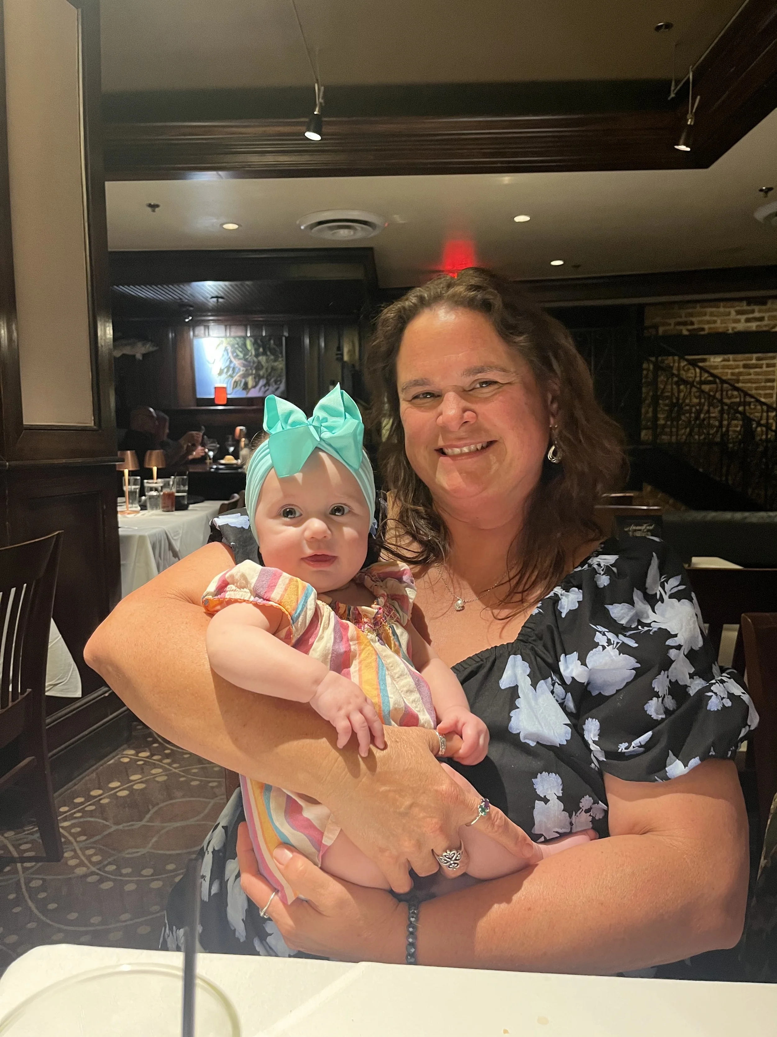 Woman holding a baby girl with a teal bow headband in a restaurant setting.
