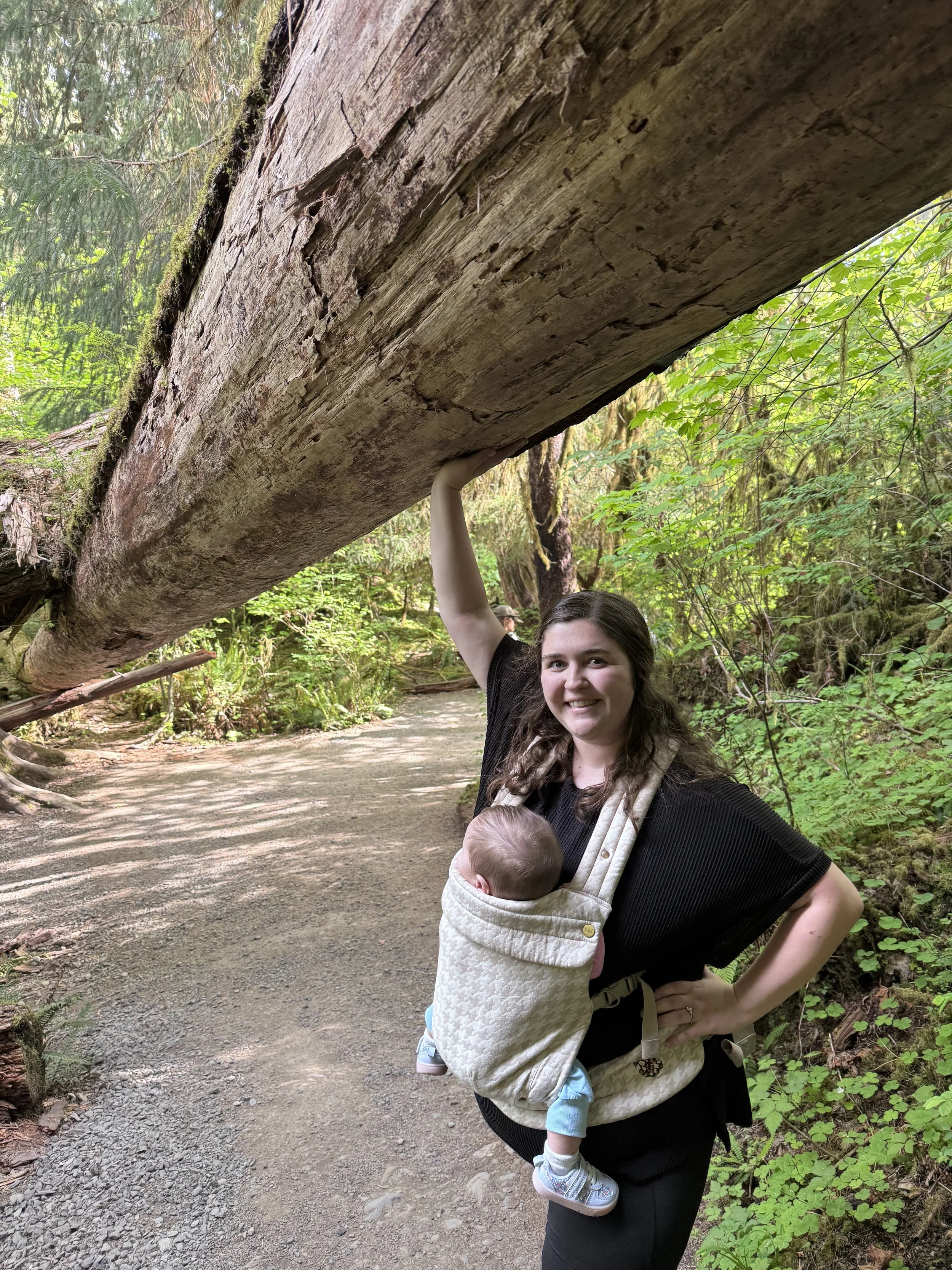 A woman with long brown hair carrying a baby in a front-facing baby carrier on a forest trail. The woman is holding a large fallen tree trunk above her head for support. The trail is surrounded by green foliage and tall trees.