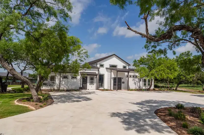 White modern house with black trim, surrounded by trees, with a large concrete driveway and landscaped areas.