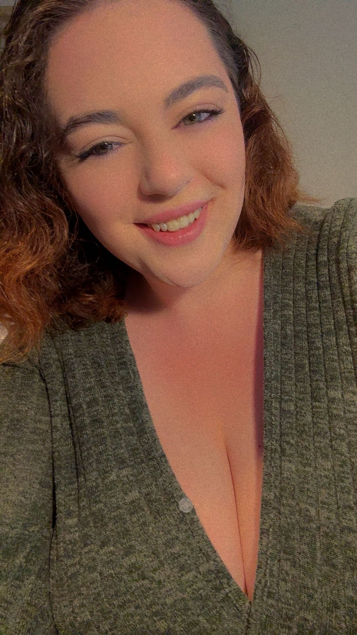 A woman with curly brown hair, light makeup, and a warm smile, wearing a gray v-neck top with subtle pinstripes, indoors with a plain background.