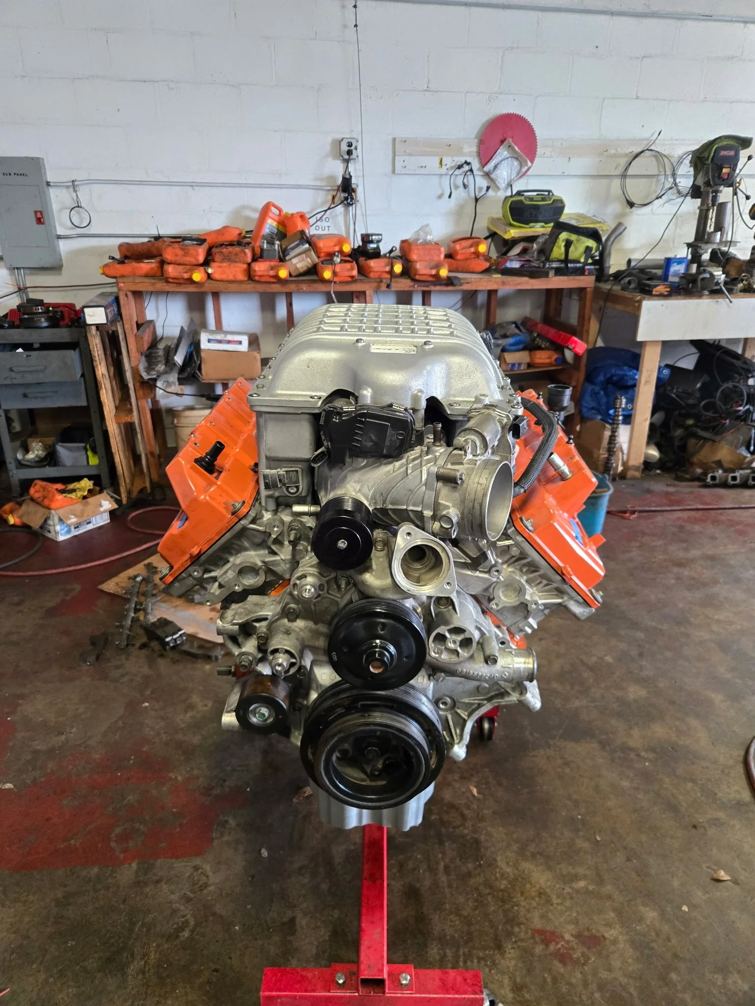 A car engine on a stand in a workshop, with various tools and supplies on shelves behind it.