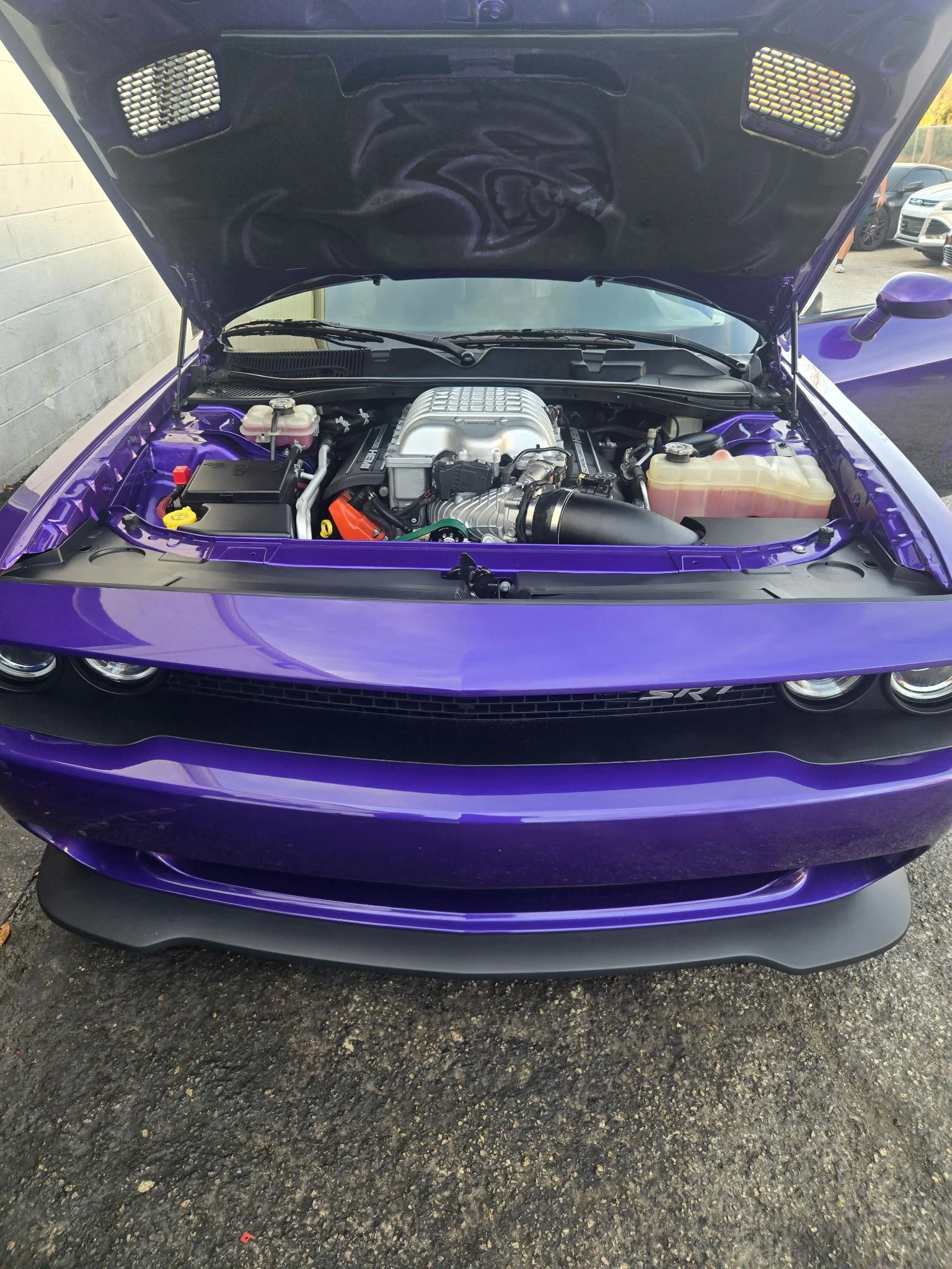 Front view of a purple muscle car with its hood open, showing the engine bay, parked on a paved surface.