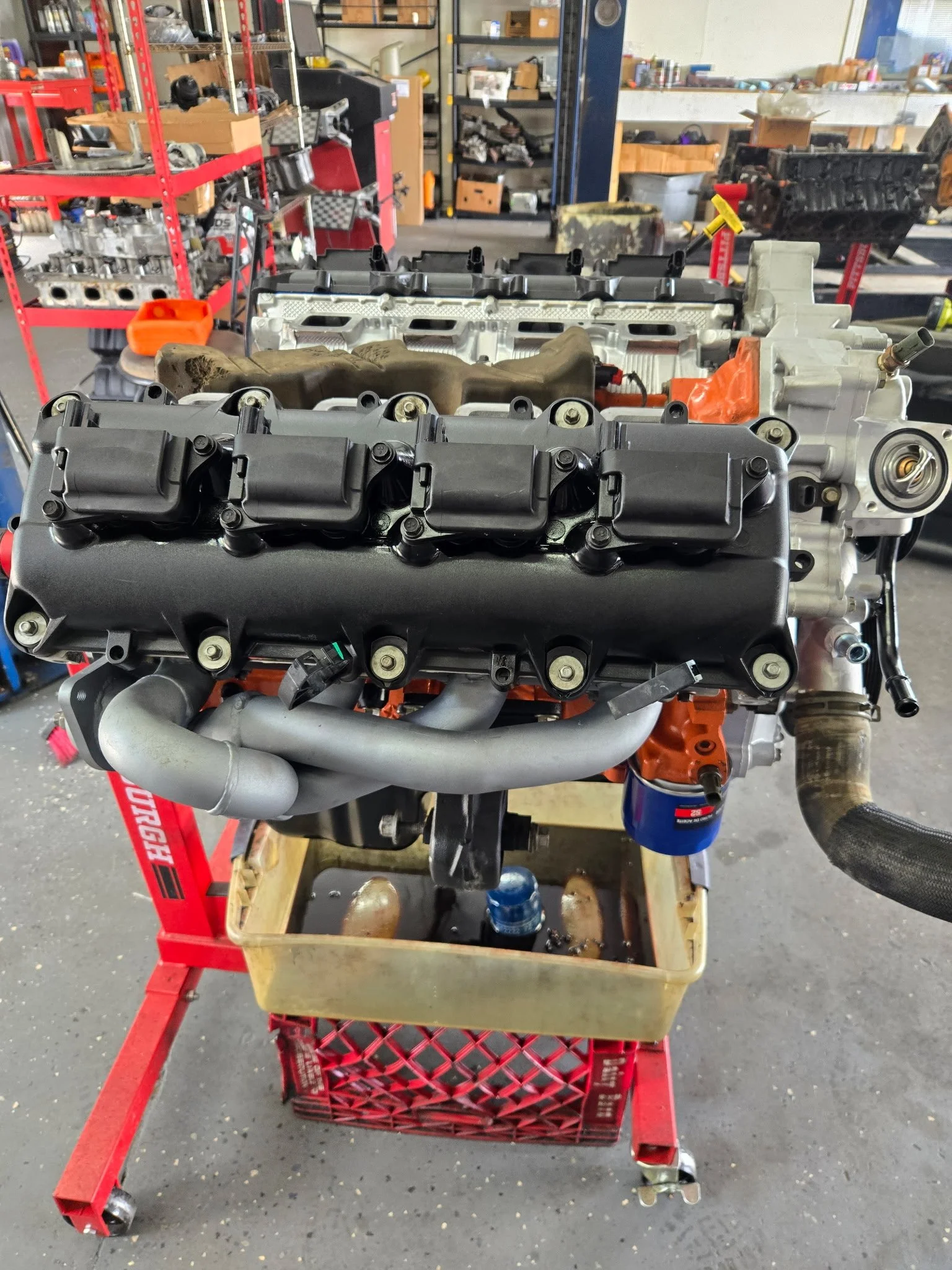 Car engine in a workshop, placed on a red metal cart with a yellow plastic bin underneath. The background features shelves with automotive parts and tools.