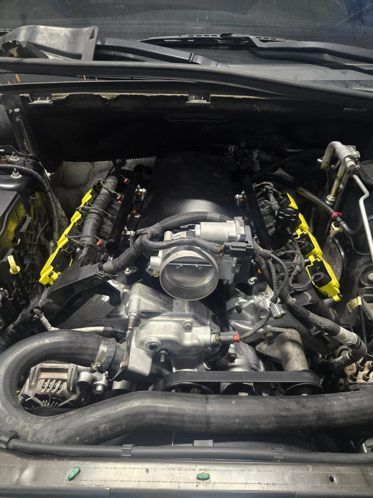 The image shows a car engine bay with the hood open, revealing various engine components, hoses, and wires. The engine appears to be a modern, high-performance type with visible yellow accents on some parts.