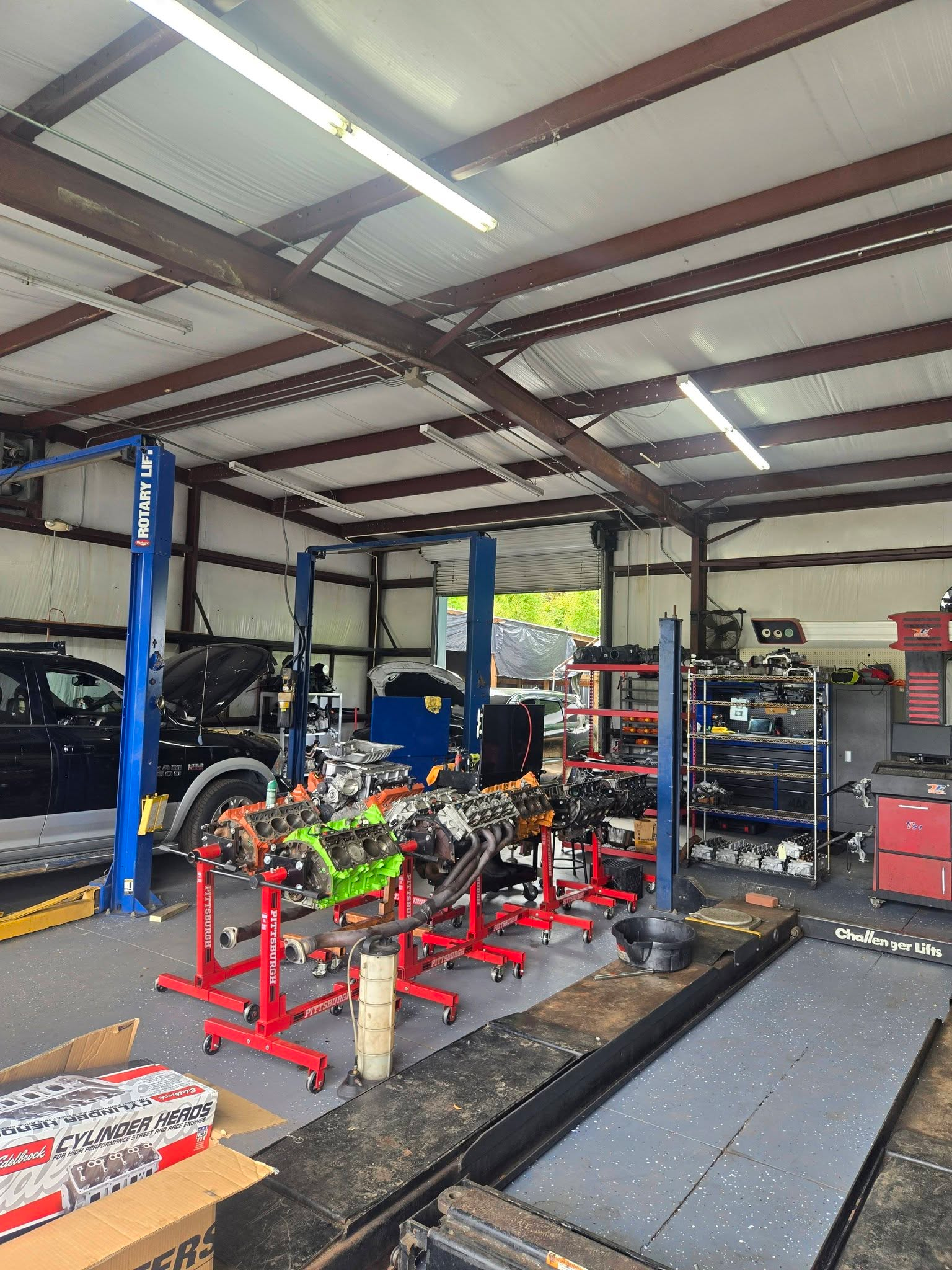 An auto repair shop interior with two cars raised on hydraulic lifts and engine parts on carts in the foreground.