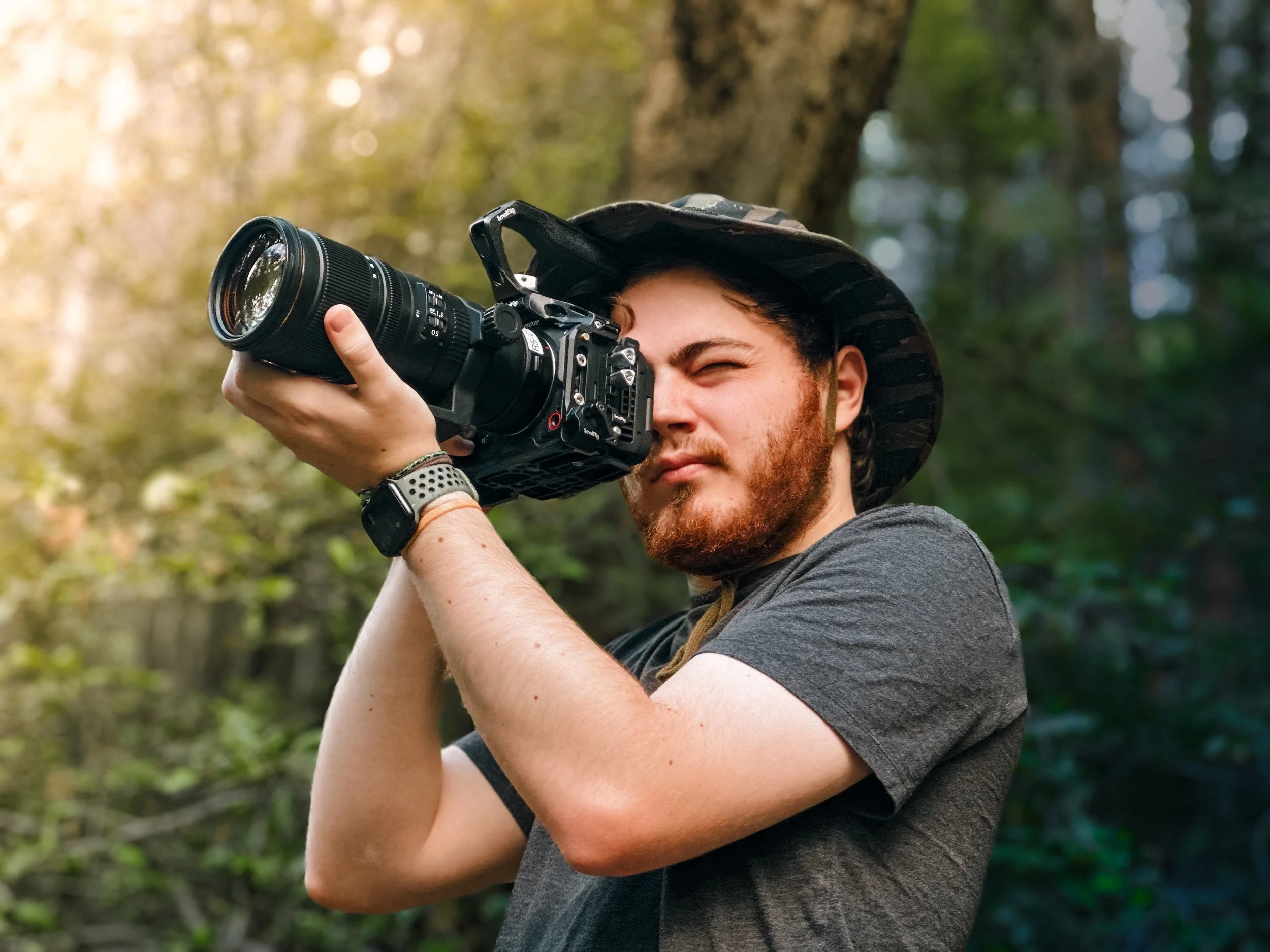 A man with a beard wearing a camo bucket hat holds a camera up to his eye, taking a photo outdoors with a background of trees and sunlight.