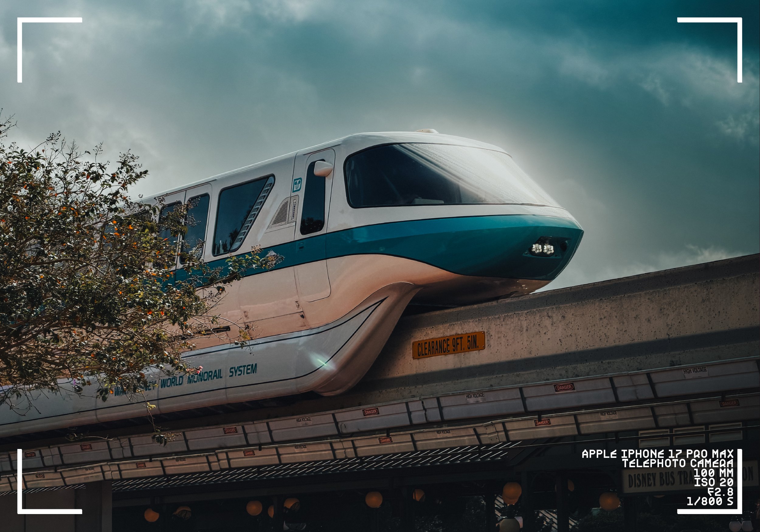 A futuristic train on an elevated track with the sky and clouds in the background. Part of the train and the track warning signs are visible, surrounded by some plants.