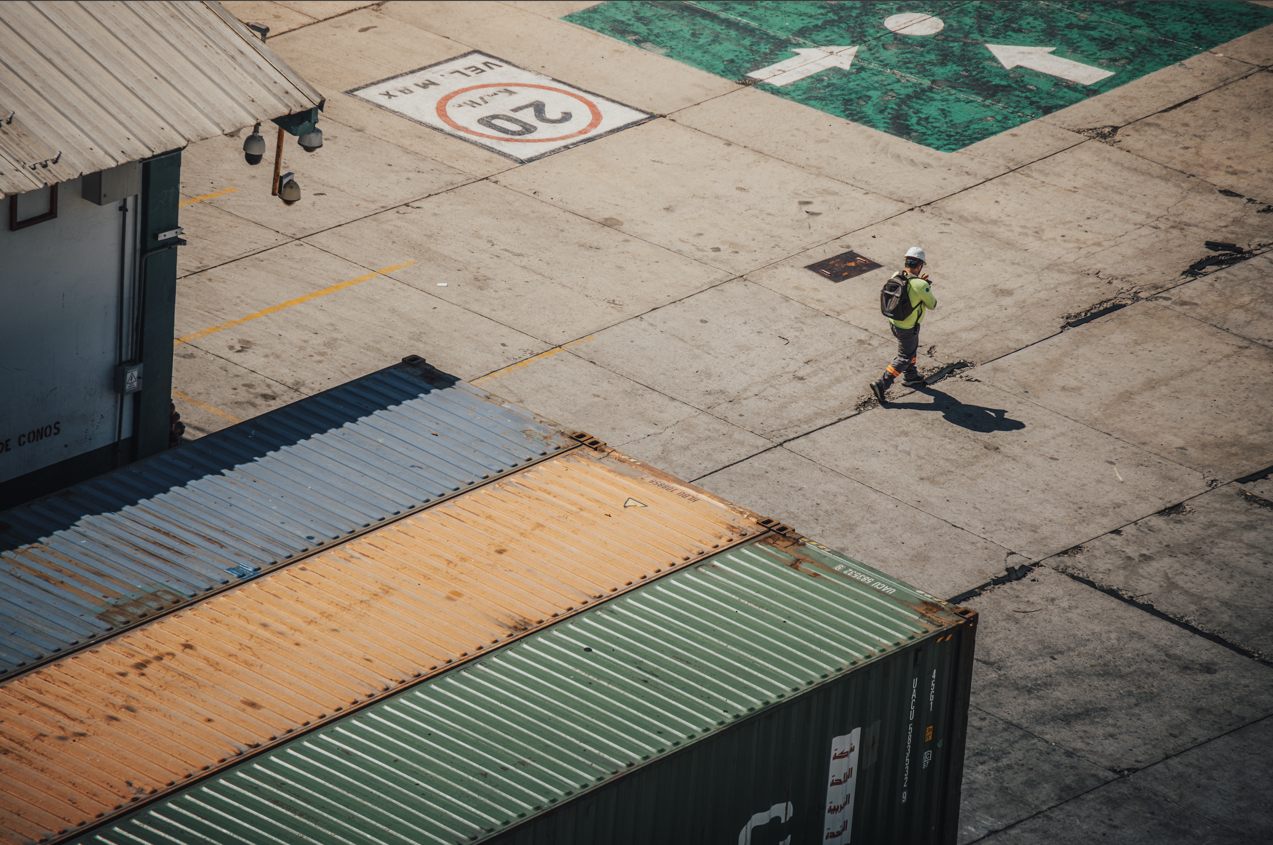Aerial view of a construction worker walking past stacked shipping containers on a concrete surface. The worker is wearing a white helmet, neon green shirt, gray pants, and has a black backpack. The ground features painted markings including an arrow and a painted speed limit of 20 miles per hour.