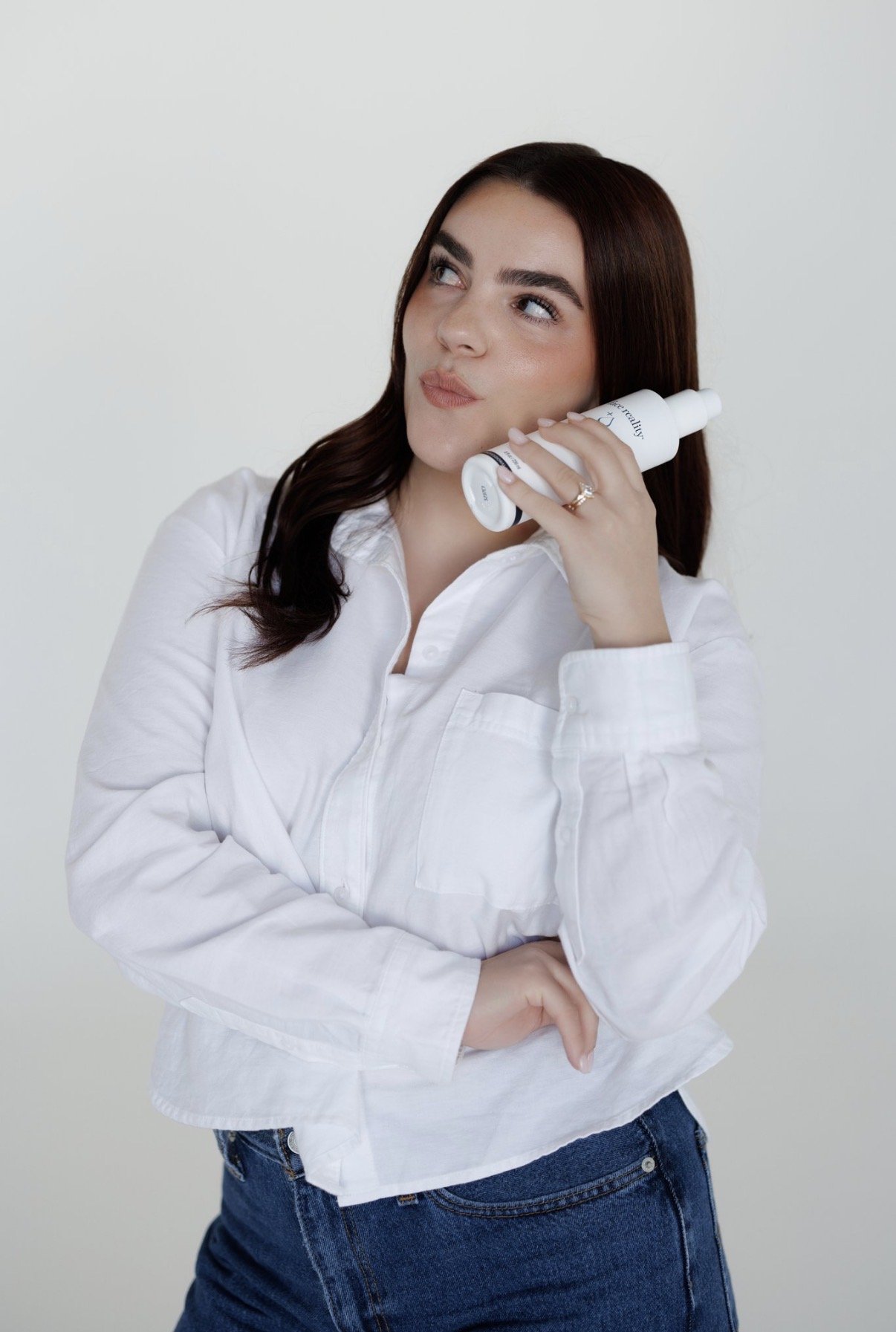 A woman with dark brown hair and light skin holds a skincare product to her face while looking to the side, against a plain light background.