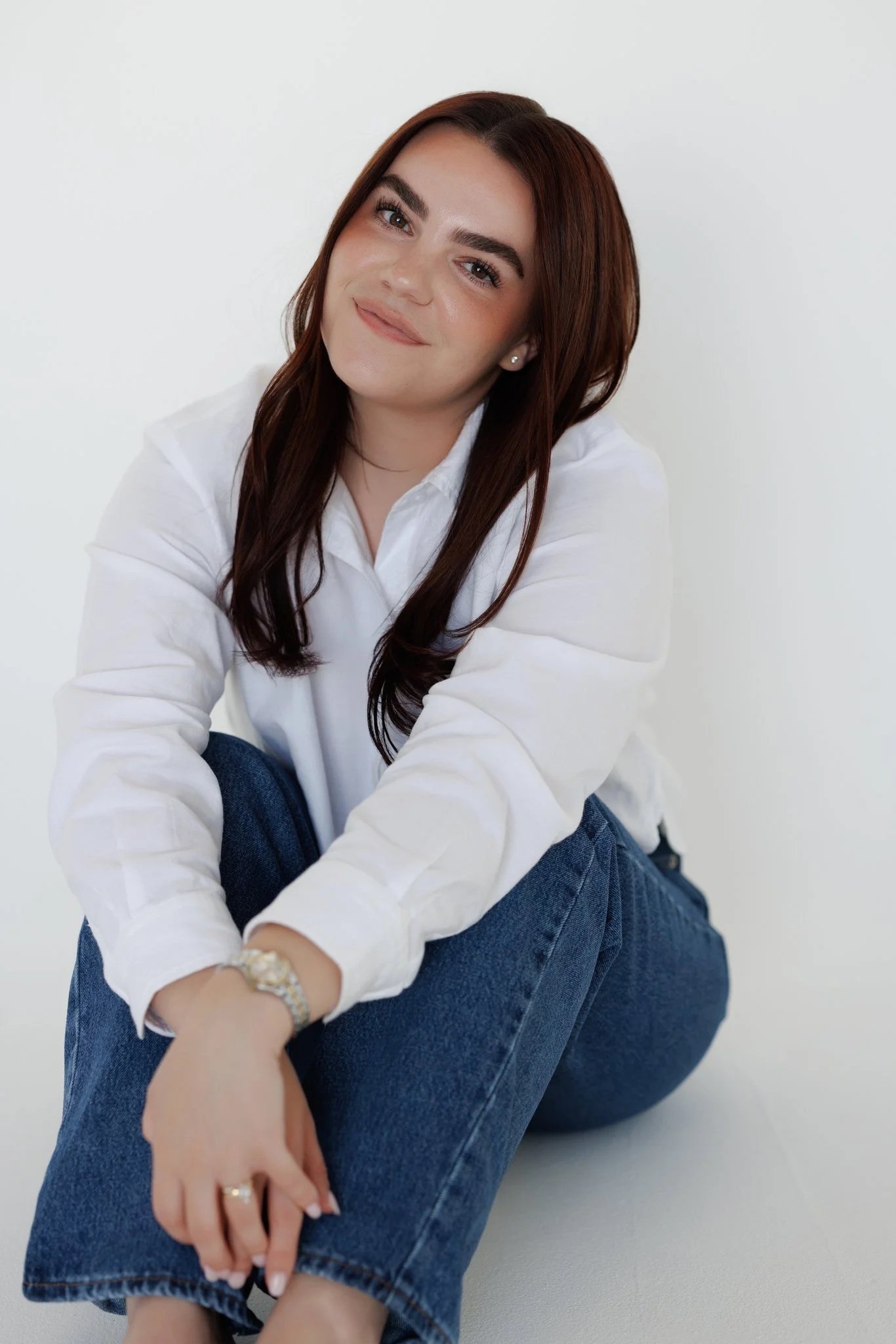 A young woman with long brown hair and earrings, wearing a white shirt and blue jeans, sitting in front of a white background.