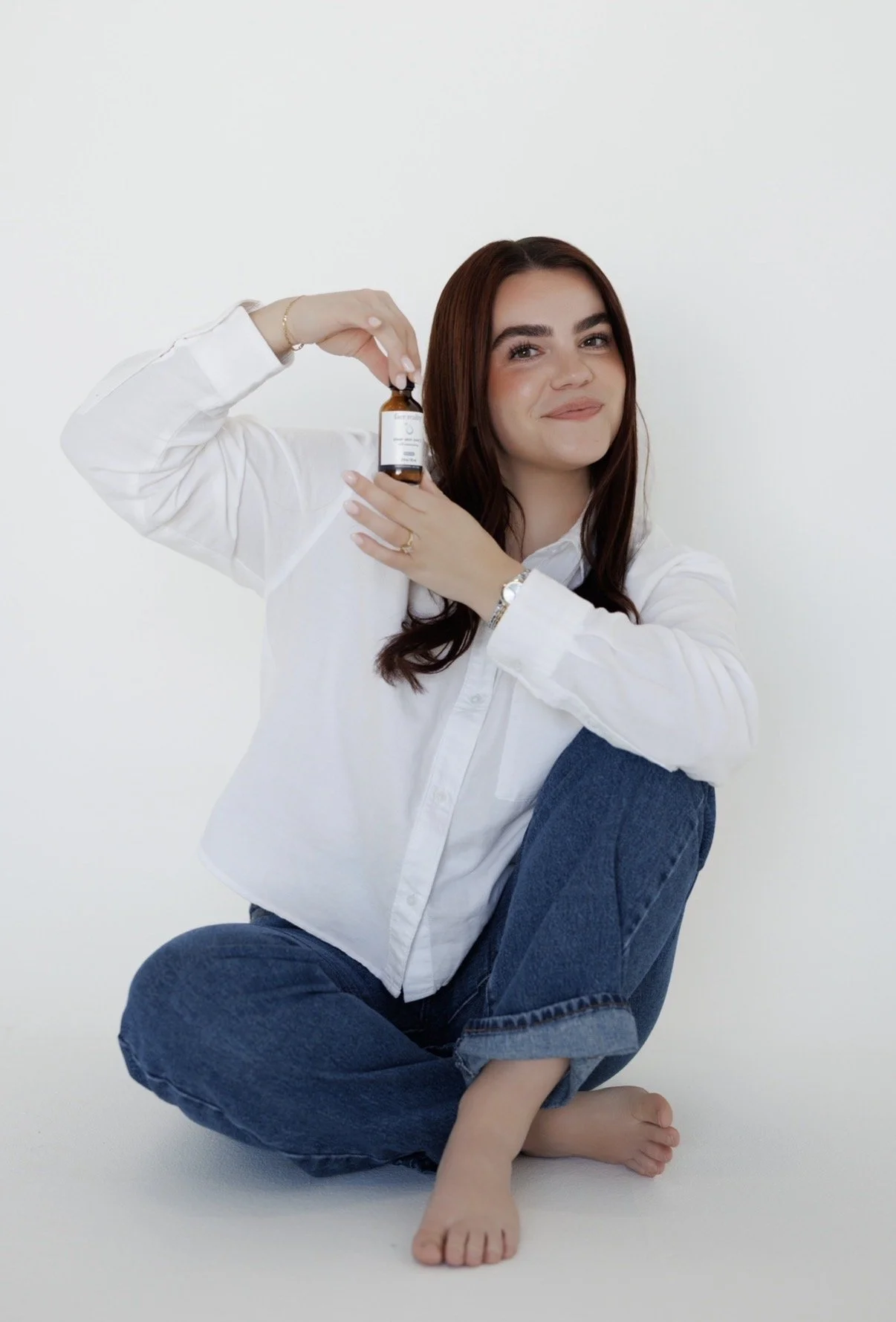 A young woman with long brown hair, wearing a white button-up shirt and blue jeans, sitting on the floor with one leg crossed under her and the other bent. She is holding a small brown bottle near her cheek and smiling softly against a plain white background.