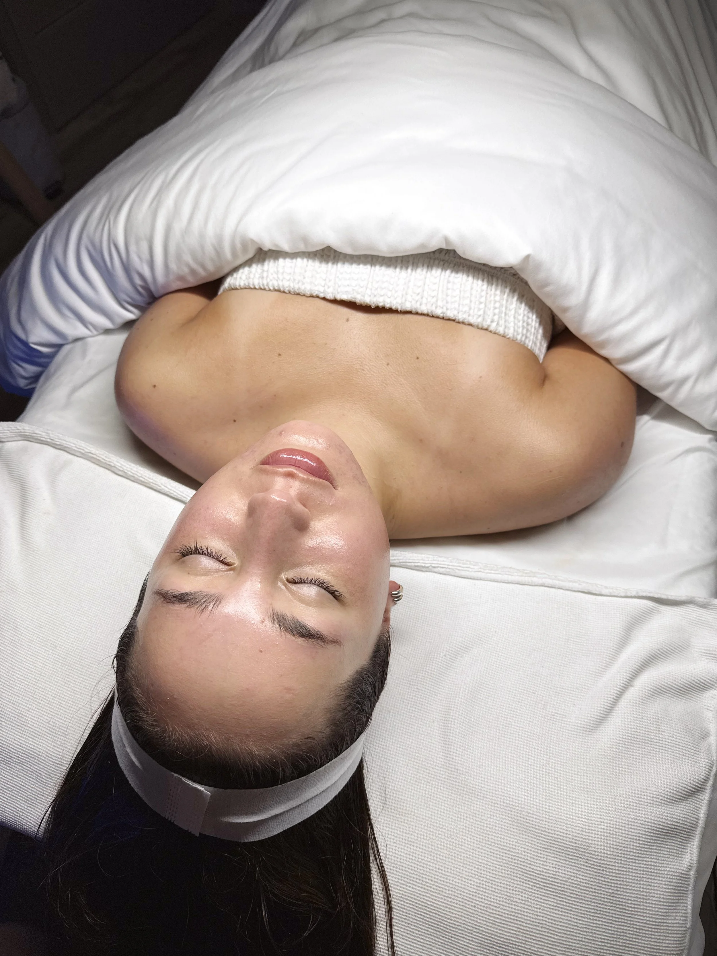 A woman lying on her back on a white bed with her eyes closed, wearing a white headband and a white tube top, covered with a white quilt.