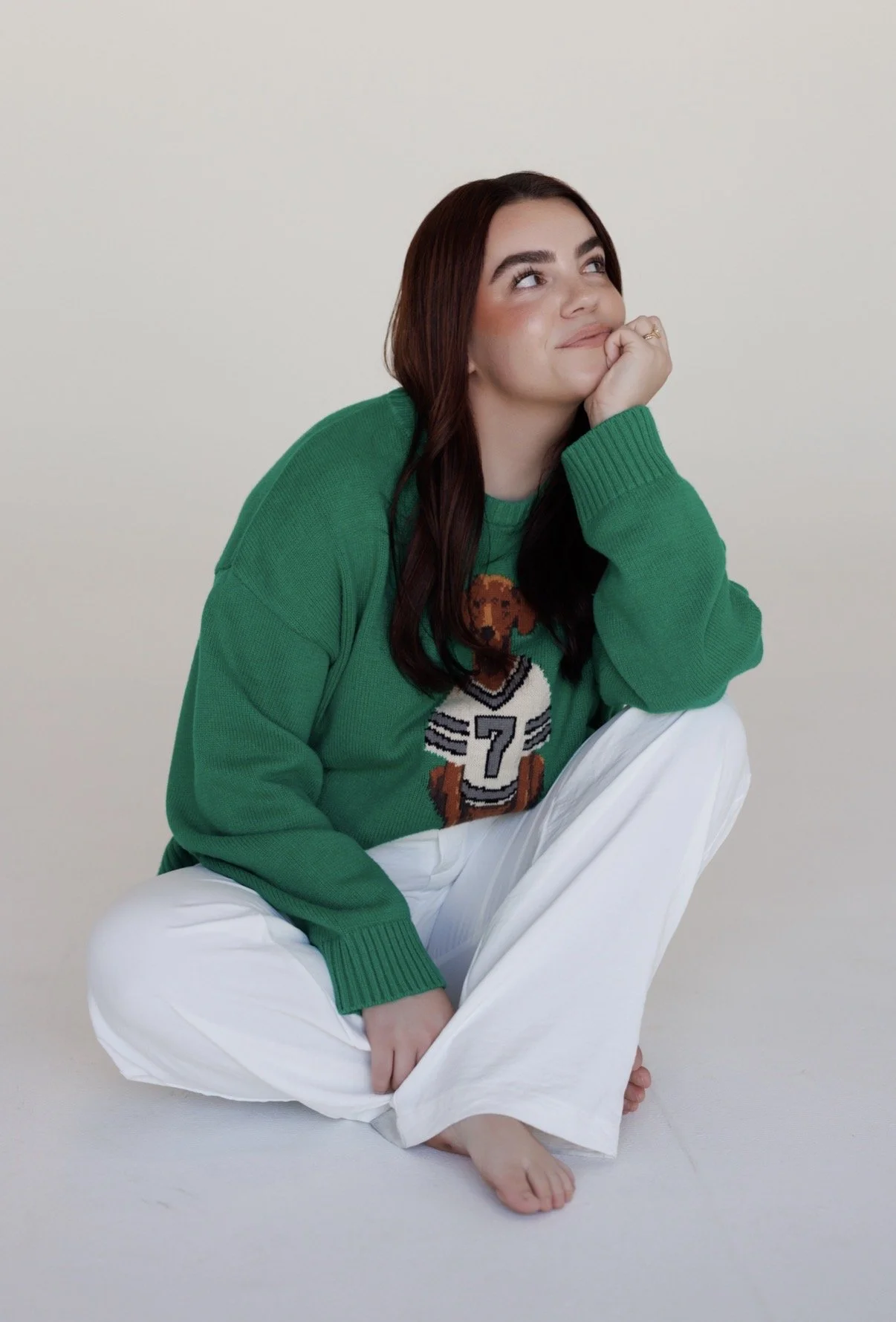 A young woman with long dark hair, wearing a green sweater with a sports design, sitting on the floor with her legs crossed and resting her chin on her hand, against a plain white background.