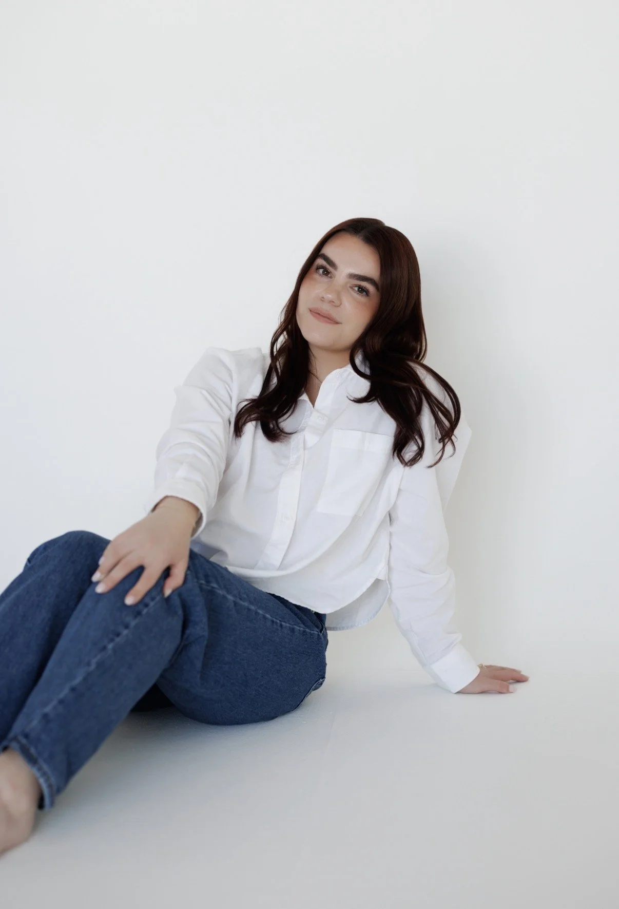 A young woman with long dark hair wearing a white shirt and blue jeans, sitting on the floor against a plain white background.