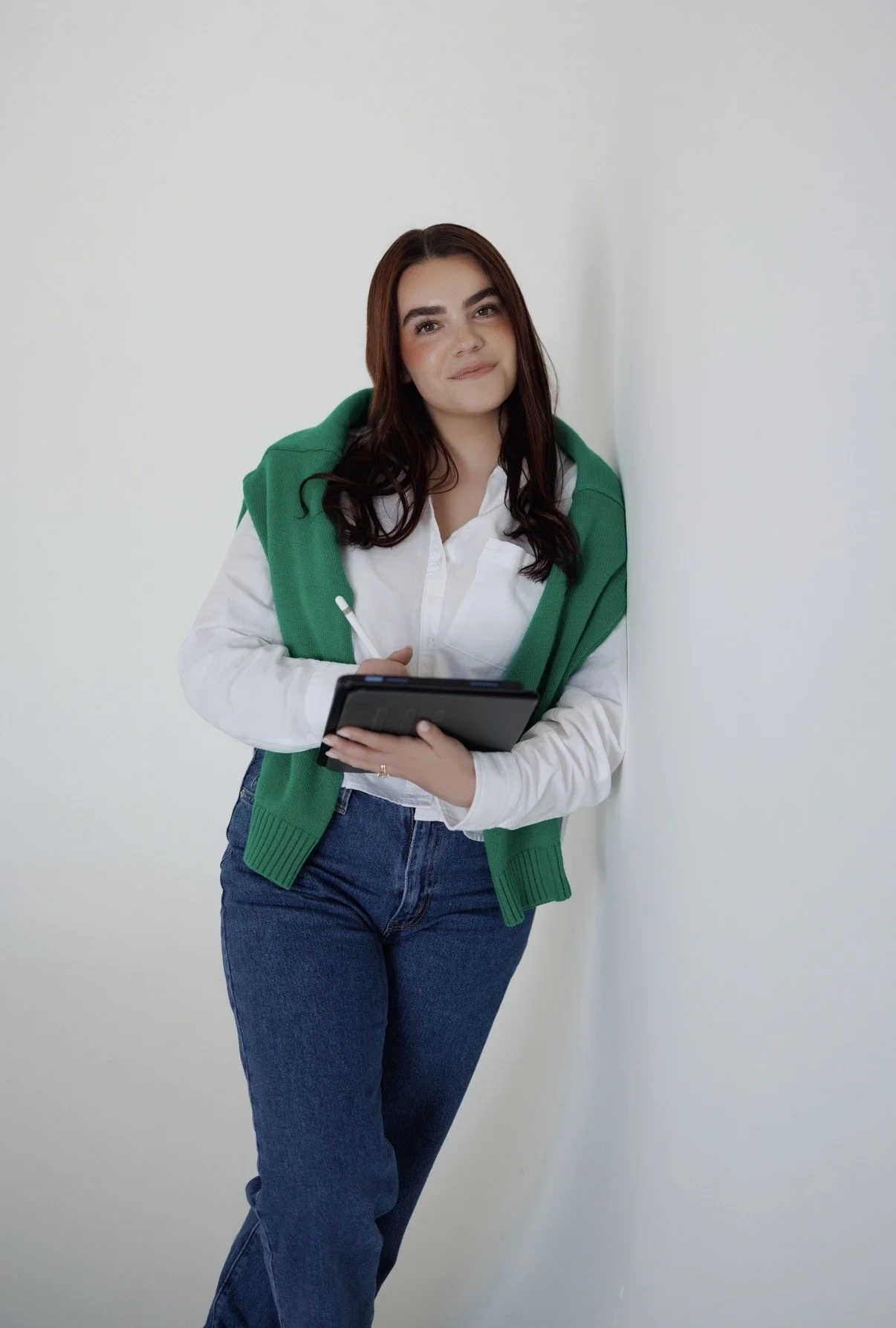 A young woman with long dark hair, wearing a white shirt and blue jeans, holding a stylus and a tablet, standing against a plain white wall with a green sweater draped over her shoulders.