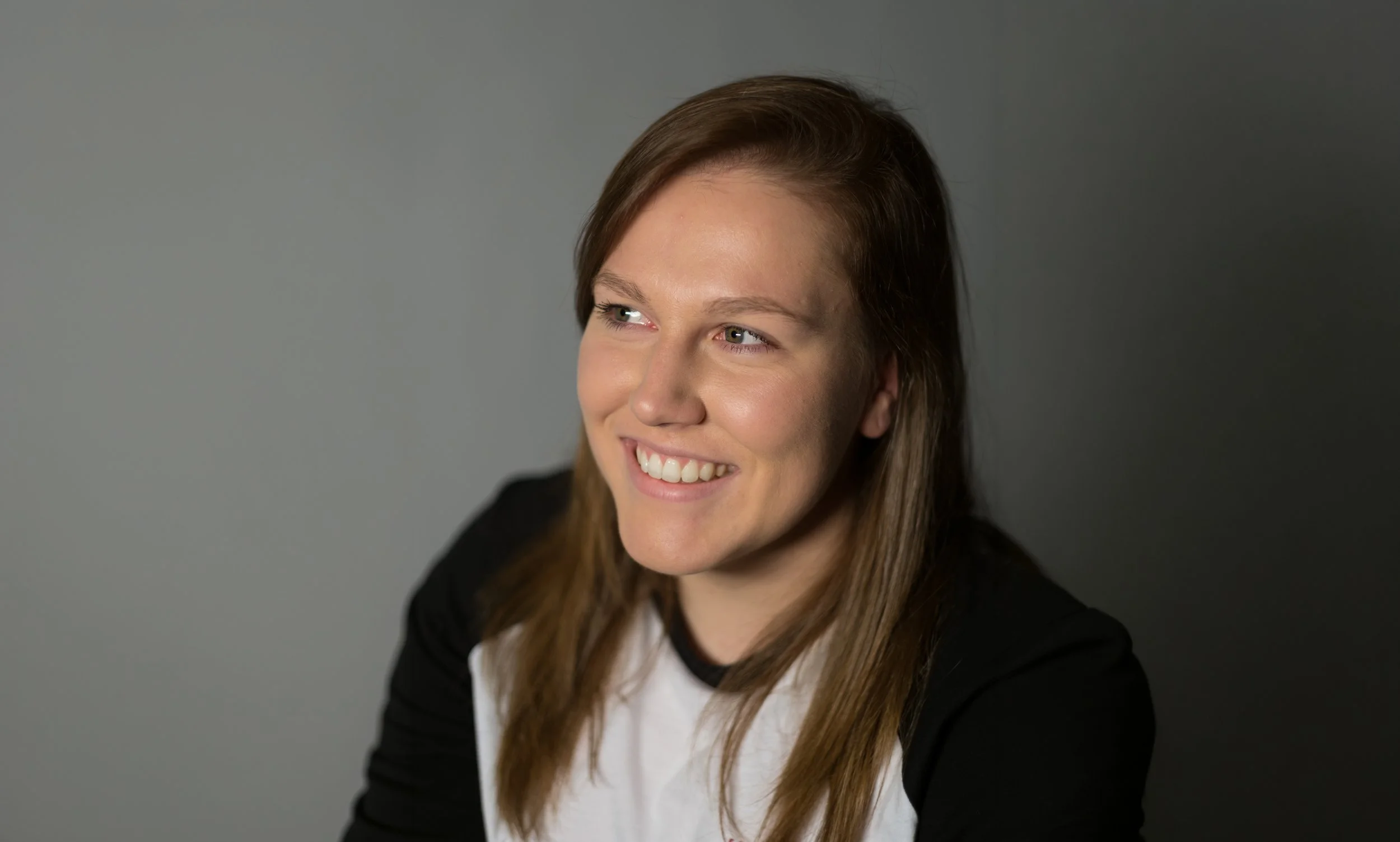 A portrait of a woman smiling and looking off into the distance, with light brown straight hair and wearing a black and white t-shirt.