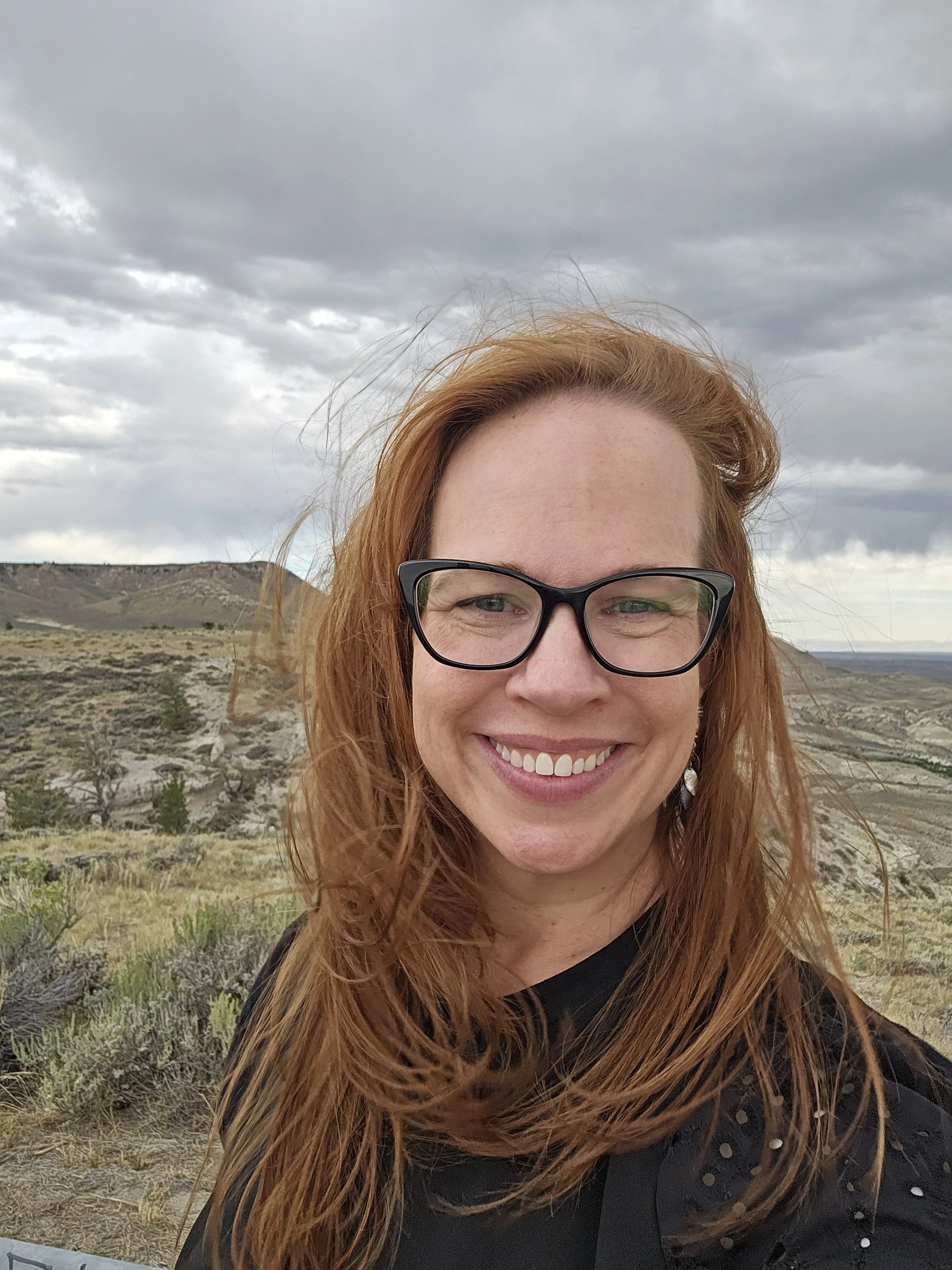 Close-up of a woman's face with red hair and black glasses, smiling outdoors with a cloudy sky and rolling hills in the background.