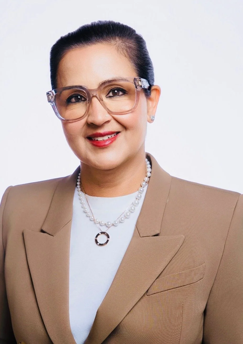 A woman wearing a beige blazer, white top, pearl necklace, and glasses, smiling against a plain white background.