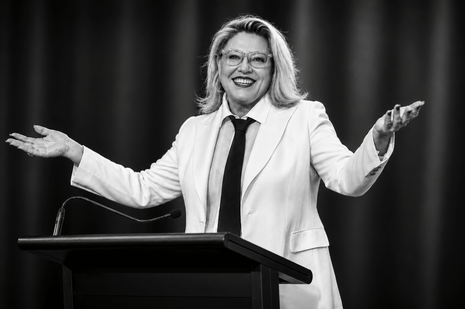 Kylie Ward wearing a white suit and collared shirt with a black tie, standing at a podium smiling with arms reaching out in a open, welcoming gesture