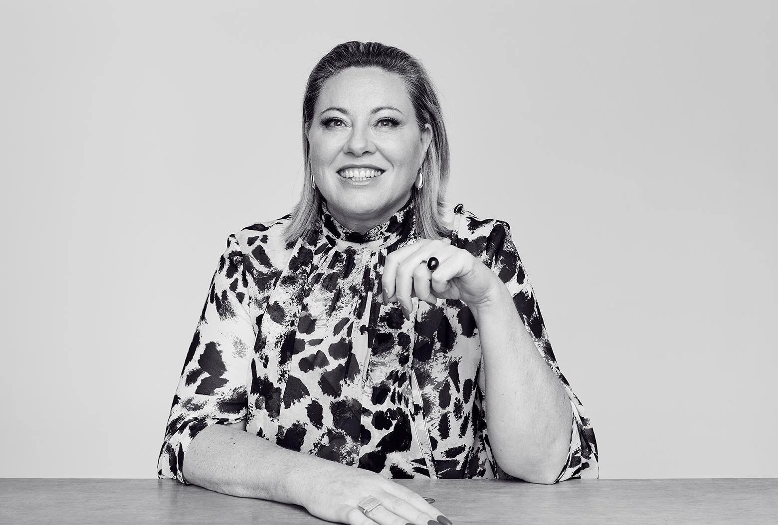 Black and white portrait of Kylie Ward with shoulder-length hair, wearing a patterned blouse with animal print, sitting at a table, smiling, with her hand resting on the table and her fingers near her chin.