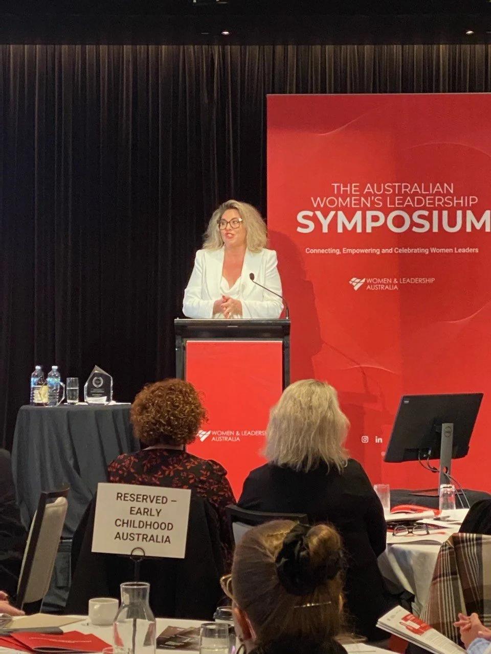 Kylie Ward speaking at a podium at the Australian Women's Leadership Symposium, with a red event banner and seated audience nearby.