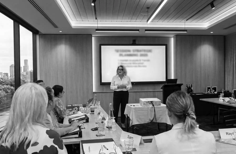 Kylie Ward presenting a presentaiton in front of a screen in a modern conference room with several attendees seated at a table.