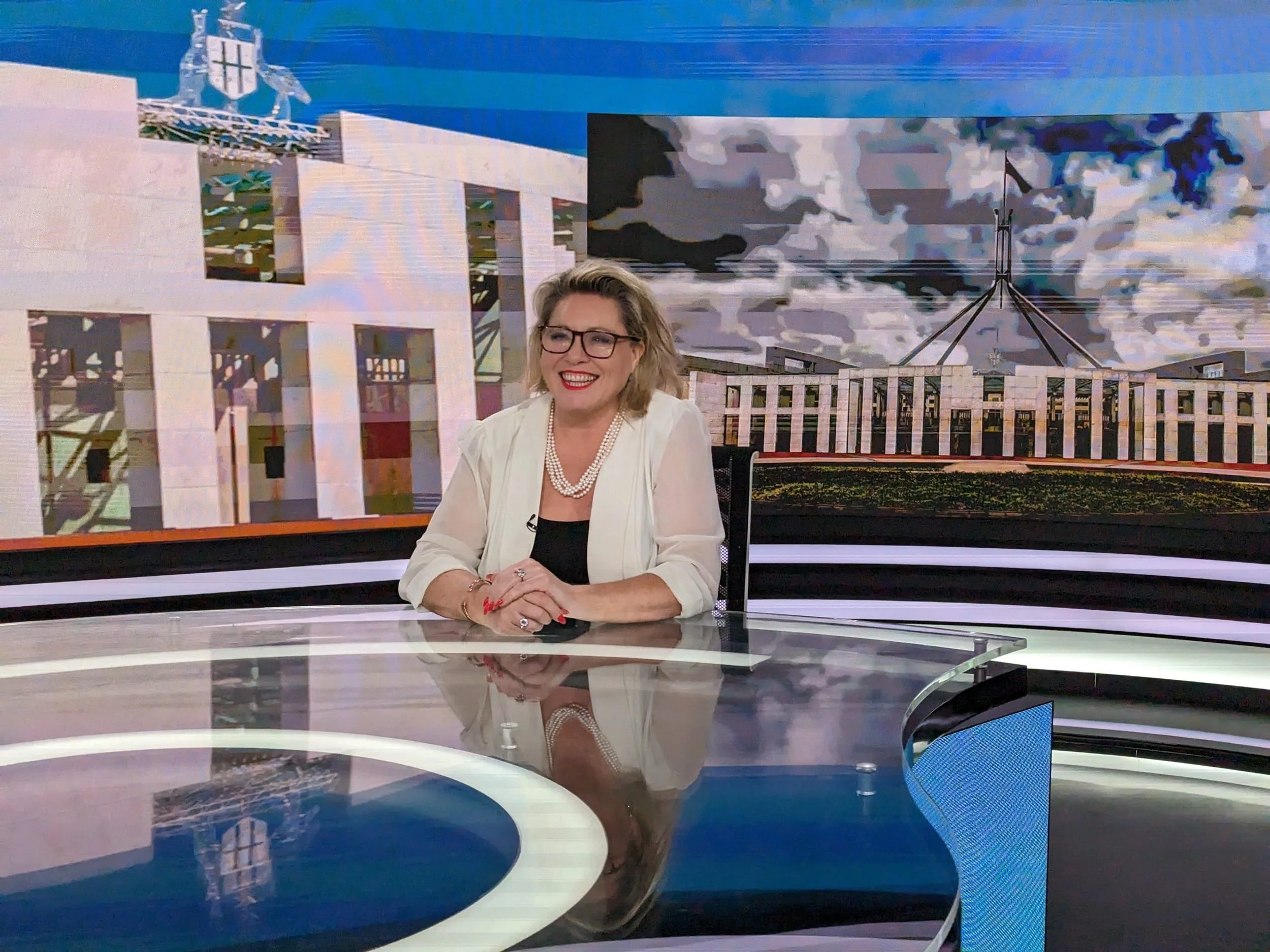 Kylie Ward seated at a television news desk, wearing a white blazer and pearls, with a Parliament House backdrop.