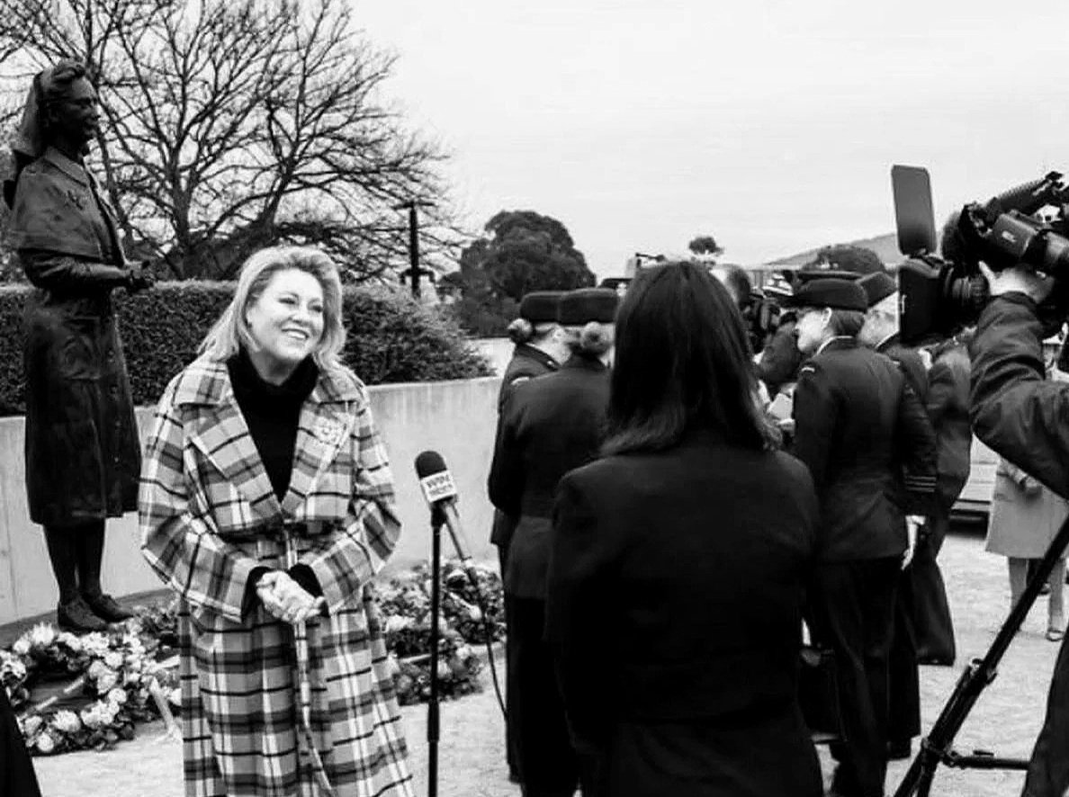 Kylie Ward in a patterned winter coats, standing outside smiling as she is interviewed by a televvision presenter in front of the Vivian Bullwinkel statue in Canberra