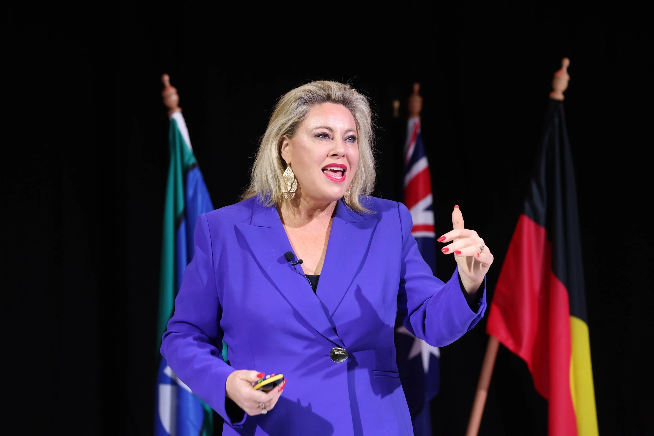 Kylie Ward speaking on stage in a purple blazer, gesturing while holding a microphone, with Australian flags in the background.