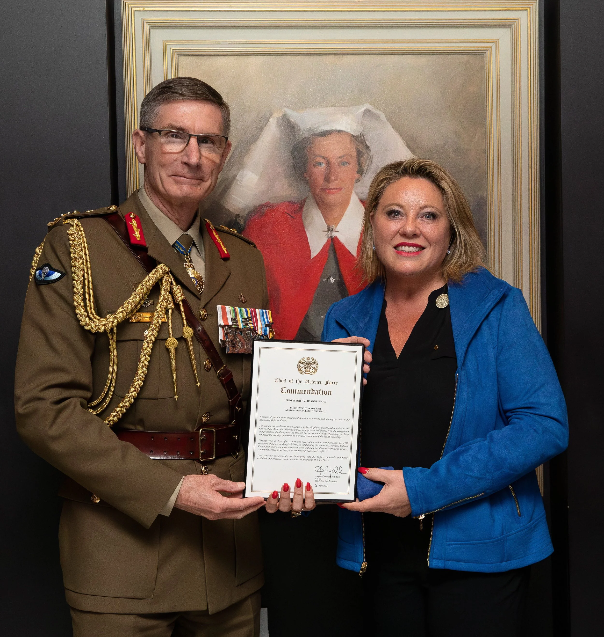 Chief of the Australian Defence Force, General Angus Campbell, in a military uniform and Kylie Ward in a blue blazer holding a certificate of Commendation together, standing in front of a portrait of celebrated nurse Vivian Bullwinkel