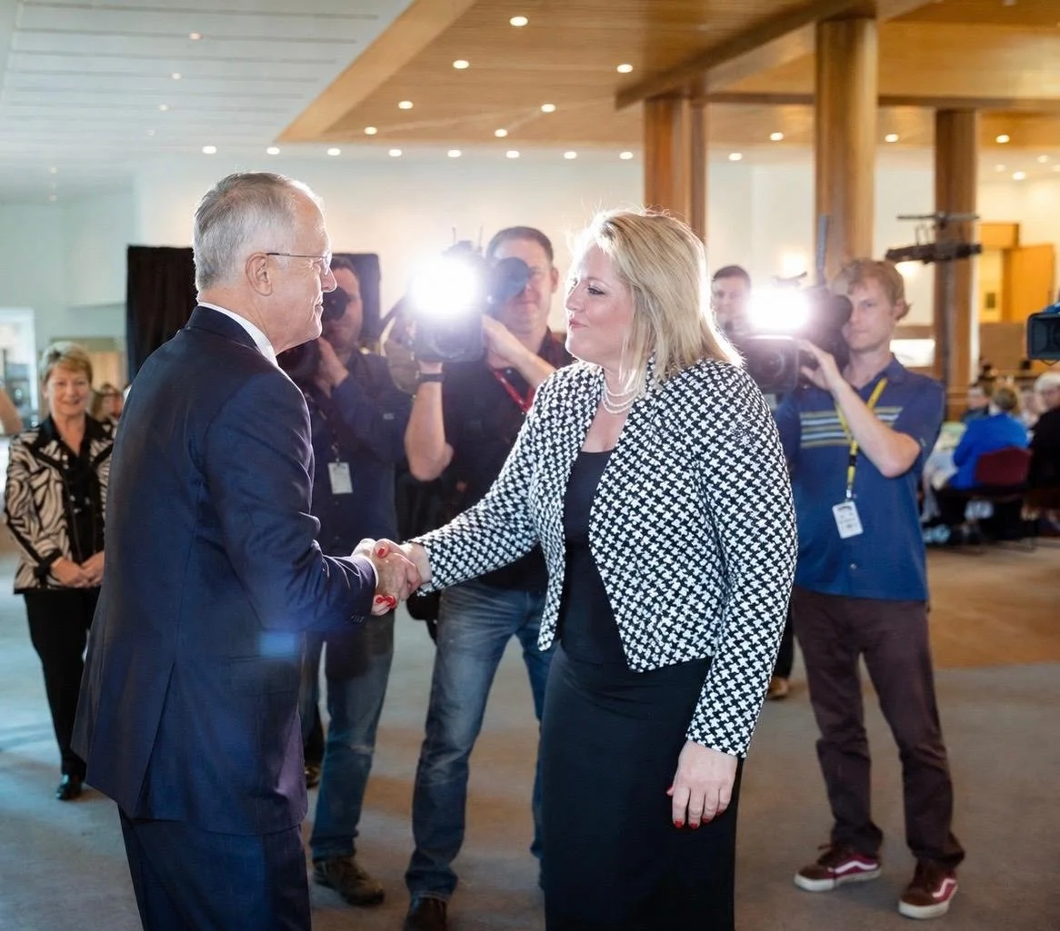 Kylie Ward and previous Australian Prime Minister Malcolm Turnbull shaking hands at an event with cameras capturing the moment, multiple onlookers in the background, and bright lighting.