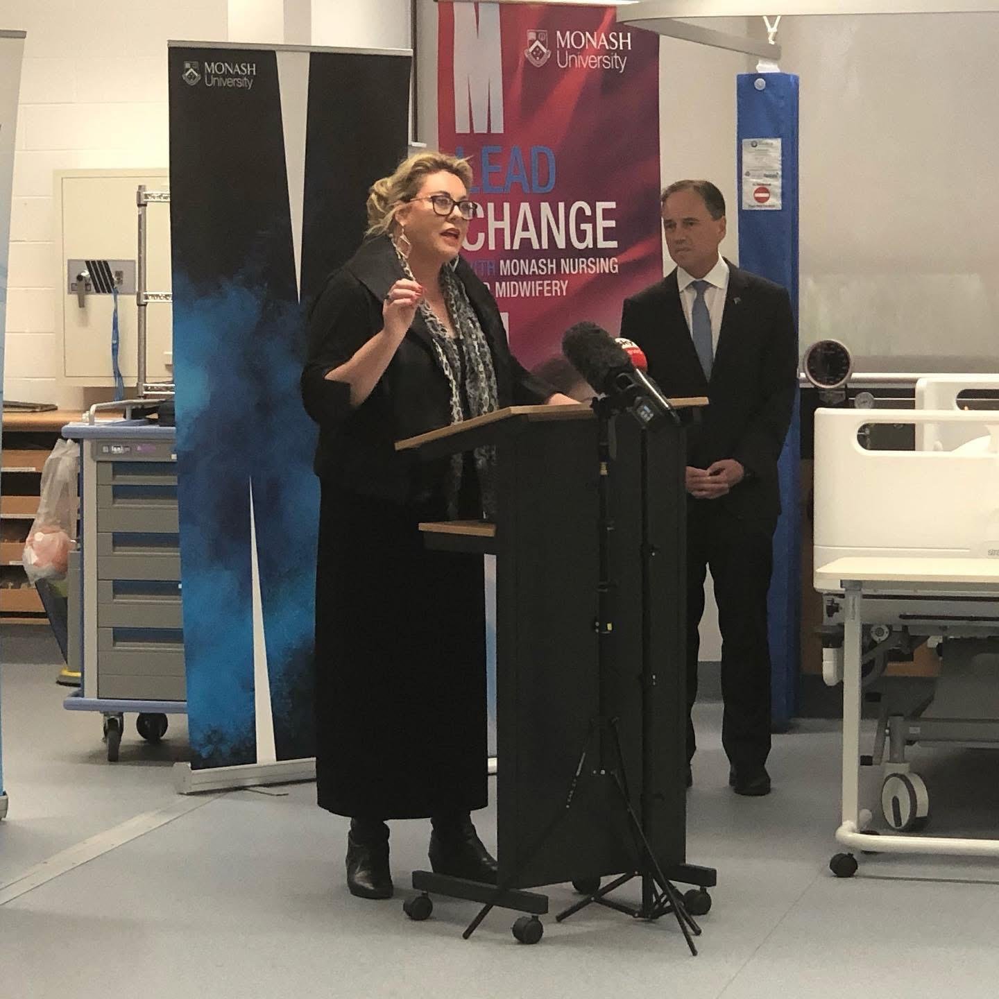 Kylie Ward speaking at a podium at a Monash University event, with a "Lead the Change" banner and a colleague standing nearby.